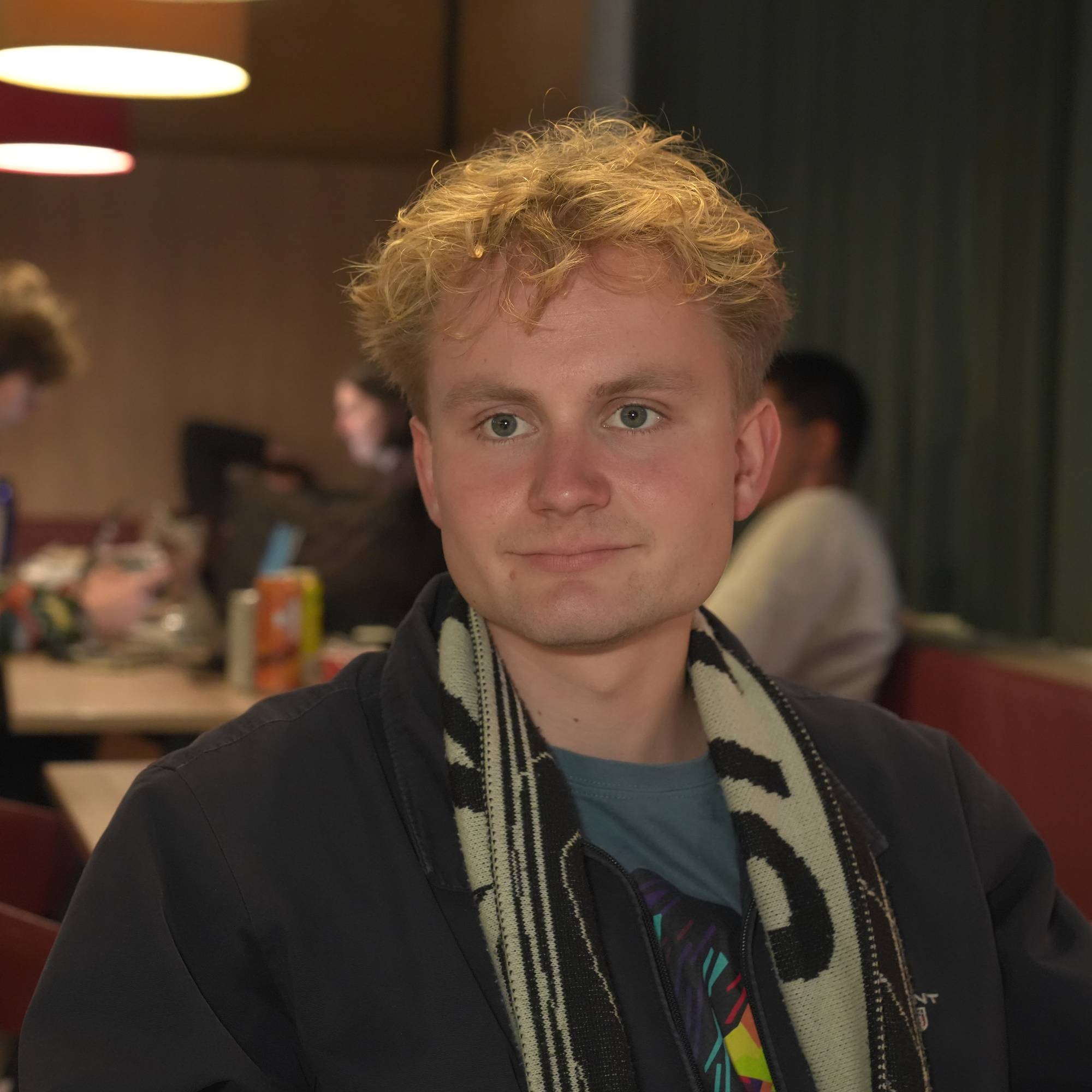 Mann med lyst krøllet hår ser i kameraet. Han har et skjerf som henger løst over skuldrene.