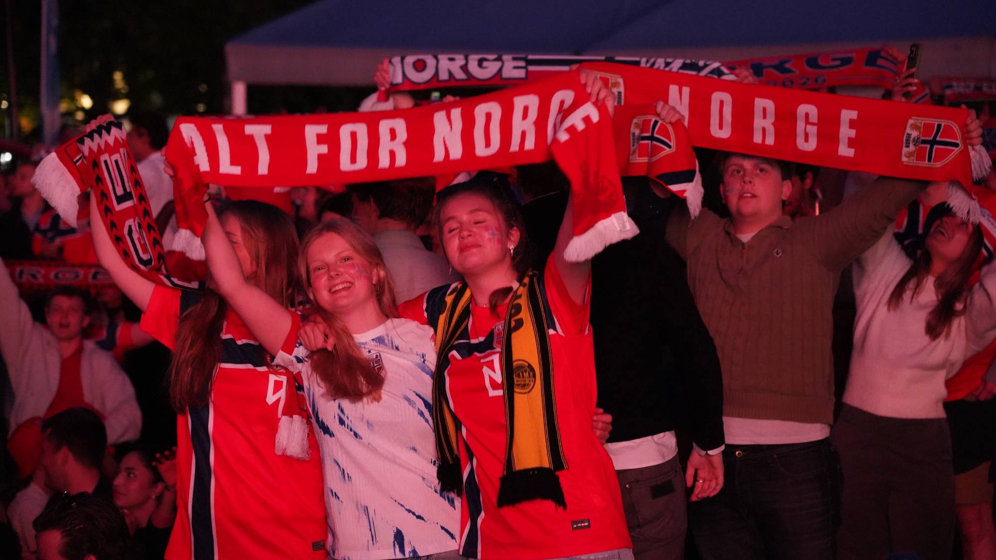 En gruppe supportere holder opp norske flagg og skjerf. Flere har malt det norske flagget i ansiktet. De står tett sammen og ser ut til å være på en utendørs arrangement. 