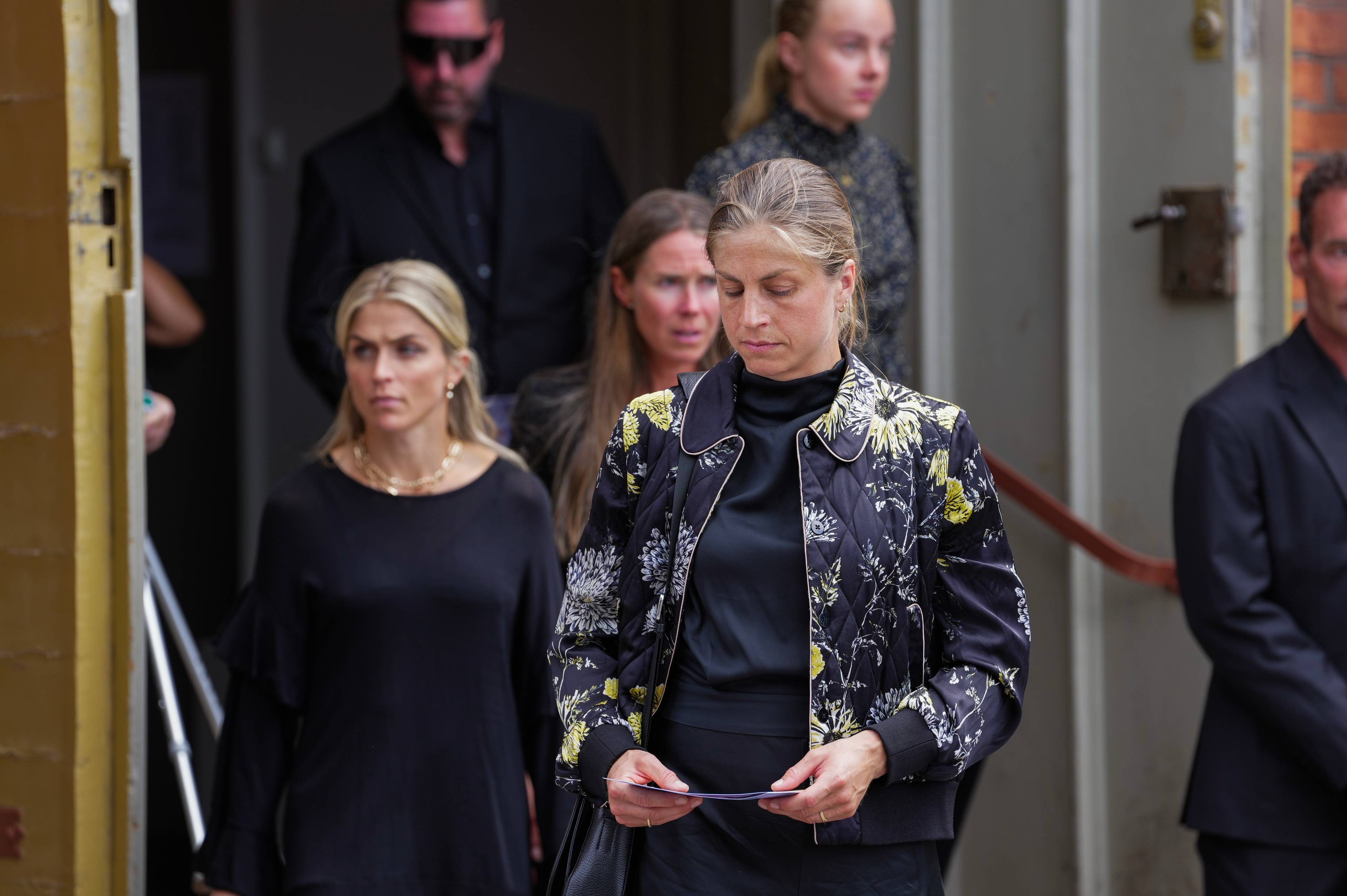Kvinner i mørke klær forlater en bygning. En kvinne i mønstret jakke holder et papir.