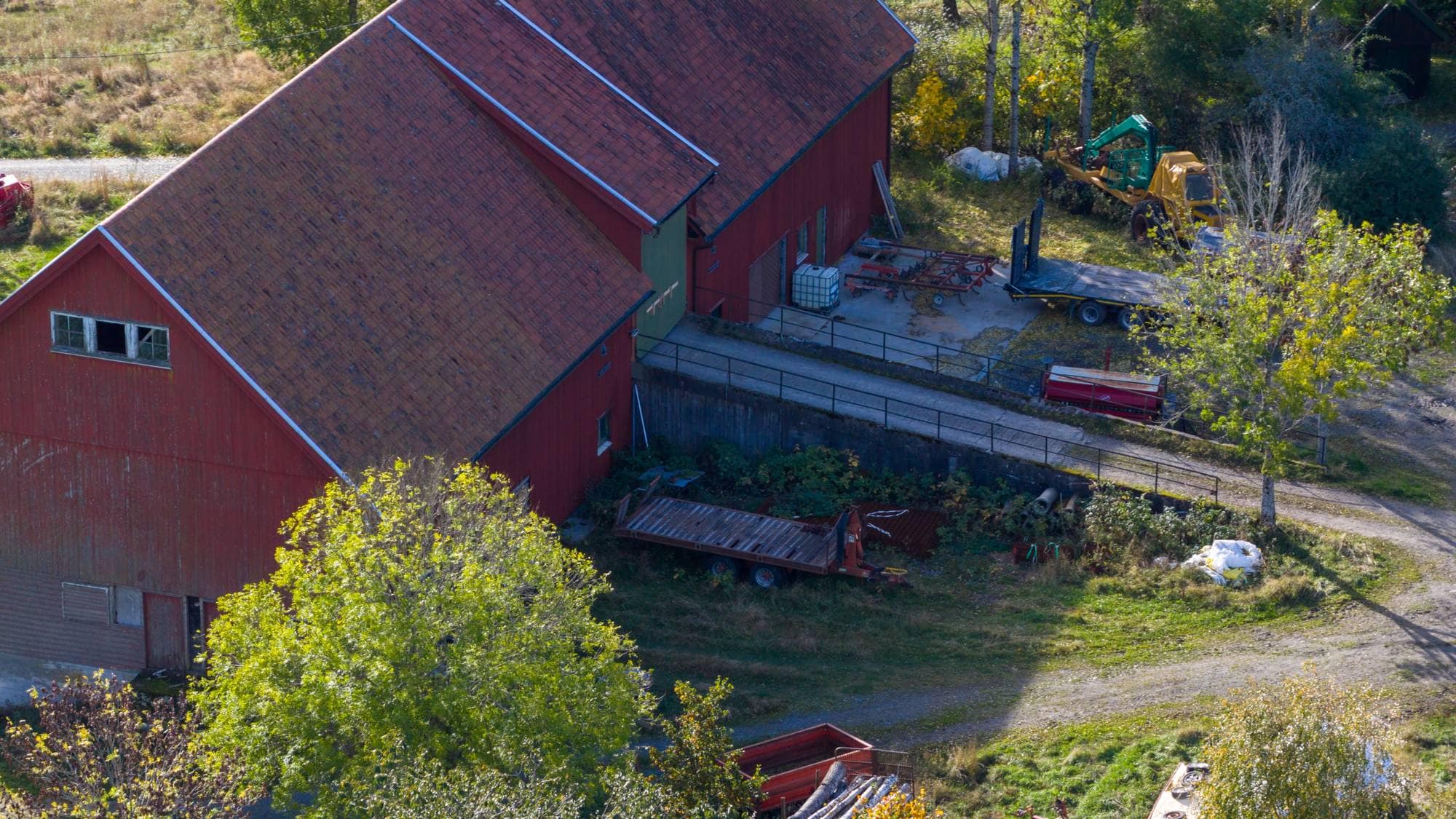 Luftfoto av en rød låve omgitt av landbruksutstyr, tilhengere og tømmerstokker på et gårdstun. (Bildebeskrivelsen er laget av en KI-tjeneste)