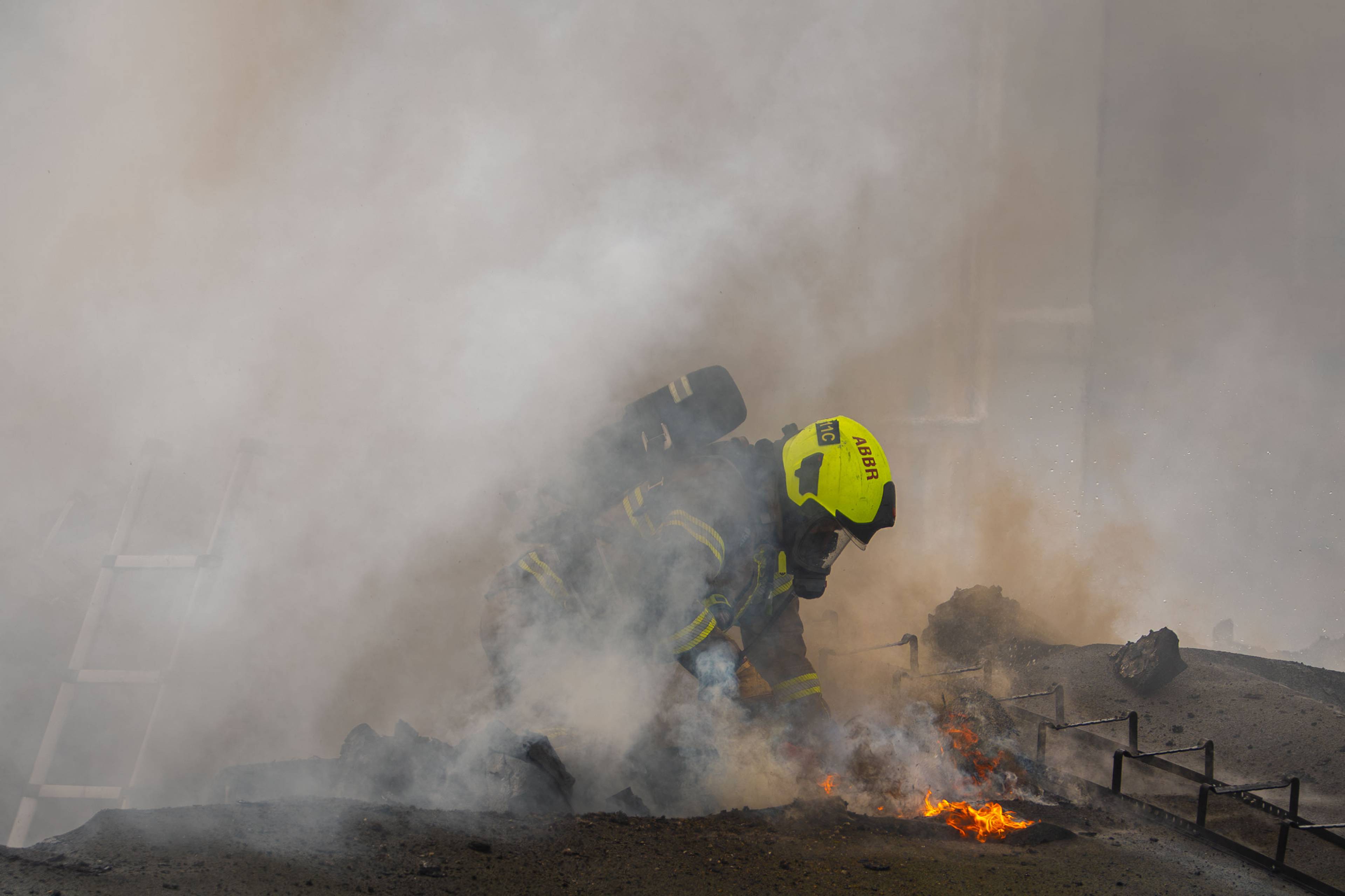 Brannmann med gul hjelm arbeider i røykfylt område. Det er flammer og stigende røyk i bildet. En stige sees til venstre. (Bildebeskrivelsen er laget av en KI-tjeneste)
