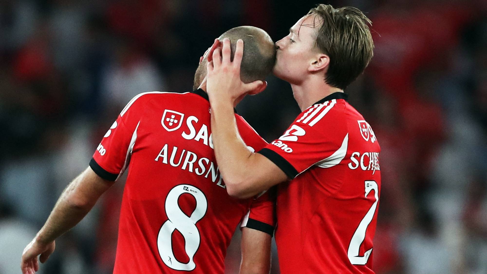 To fotballspillere i røde drakter med hvite detaljer. Den ene spilleren kysser den andre på hodet. Bakgrunnen er utydelig med røde og hvite detaljer. (Bildebeskrivelsen er laget av en KI-tjeneste)