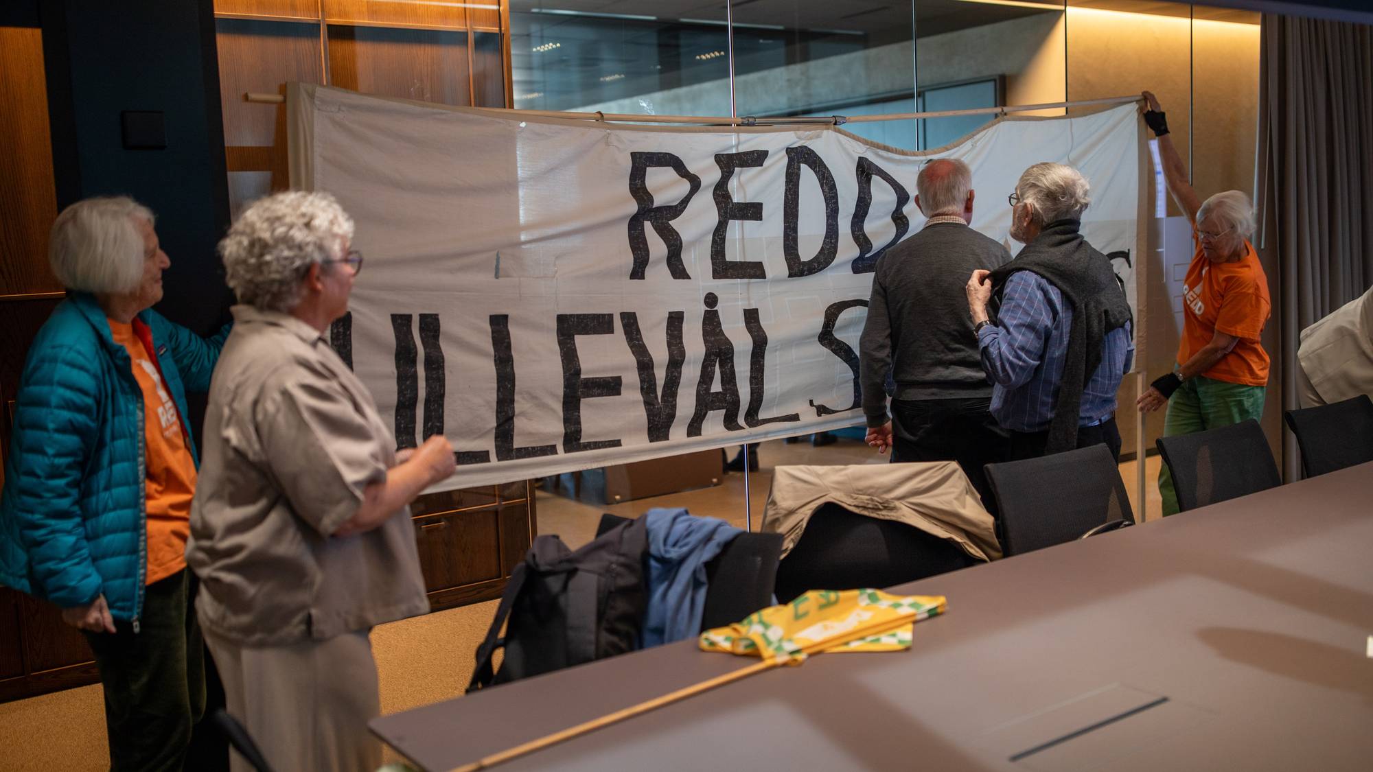 En gruppe eldre personer henger et banner i et møterom. Banneret har teksten "REDD LIVET SITT". En kvinne i oransje t-skjorte er synlig. (Bildebeskrivelsen er laget av en KI-tjeneste)