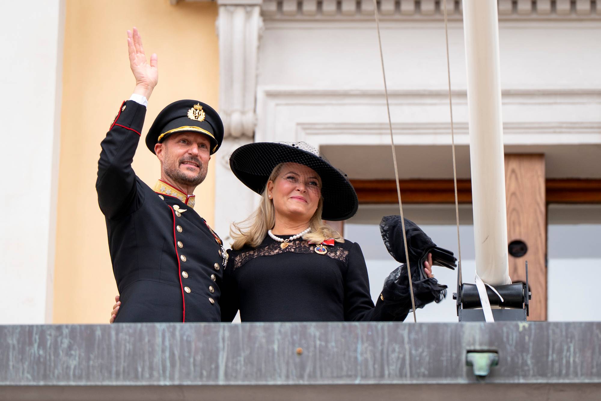 Kong Haakon vinker fra slottsbalkongen sammen med dronning Mette-Marit. De er ikledd sørgedrakter, men smiler til de oppmøtte. 