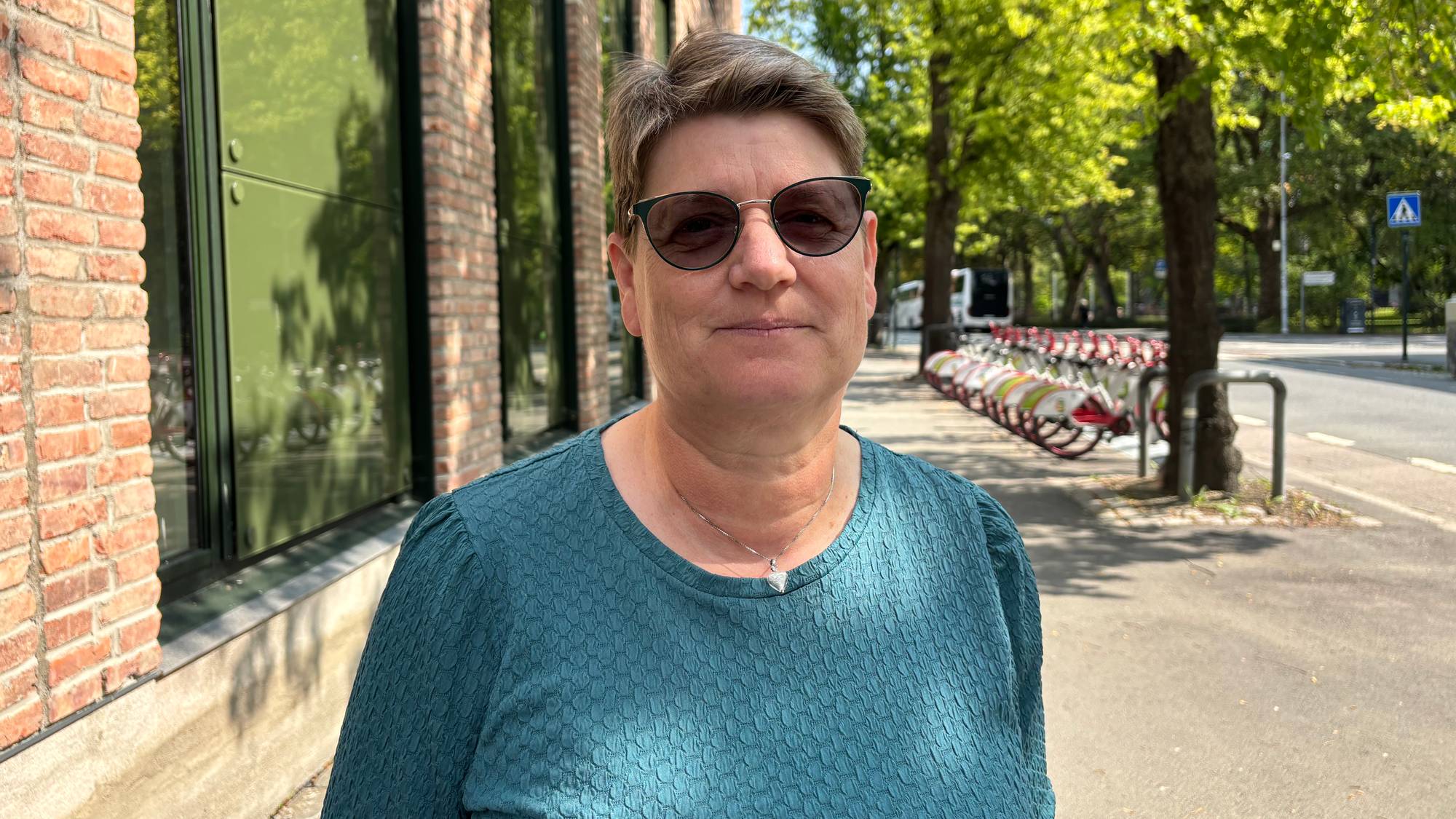 Ei dame med kort hår og solbriller står foran en murvegg med store vinduer