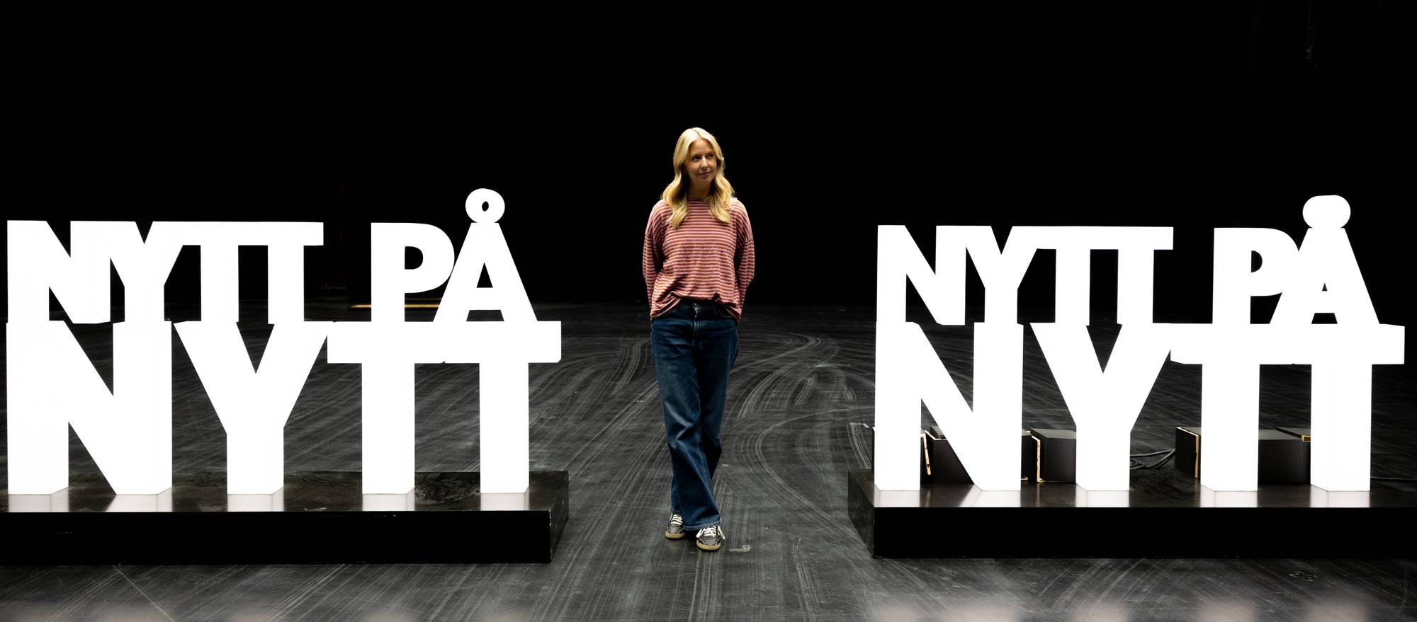 Kristine Grændsen mellom to store, hvite bokstavinstallasjoner som danner ordene "NYTT PÅ NYTT". Hun har på seg en stripete genser og jeans. Bak henne er en mørk bakgrunn. (Bildebeskrivelsen er laget av en KI-tjeneste)