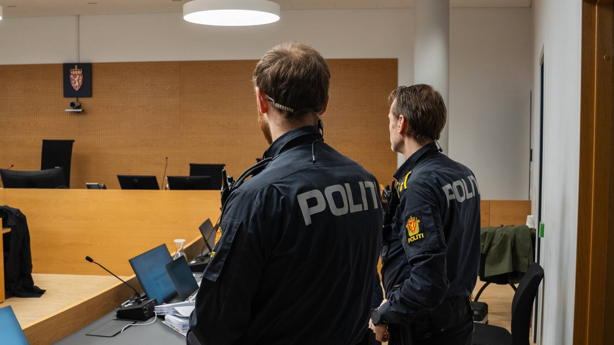To politibetjenter i mørke uniformer står i en rettssal. De har ryggen til kameraet. I bakgrunnen er et dommerpanel og et sete. (Bildebeskrivelsen er laget av en KI-tjeneste)