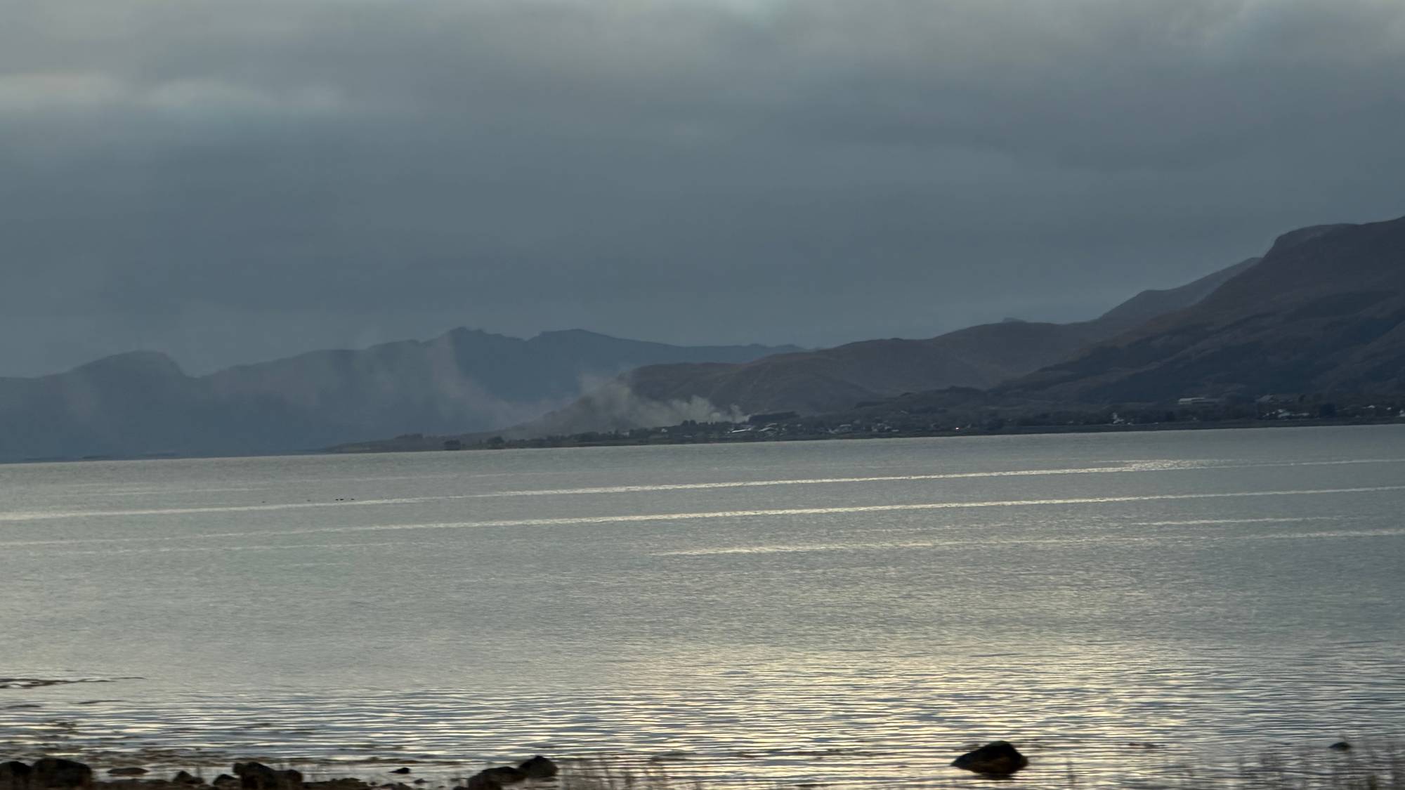 Røyk stiger opp fra et område ved en fjord med fjell i bakgrunnen under en overskyet himmel. (Bildebeskrivelsen er laget av en KI-tjeneste)