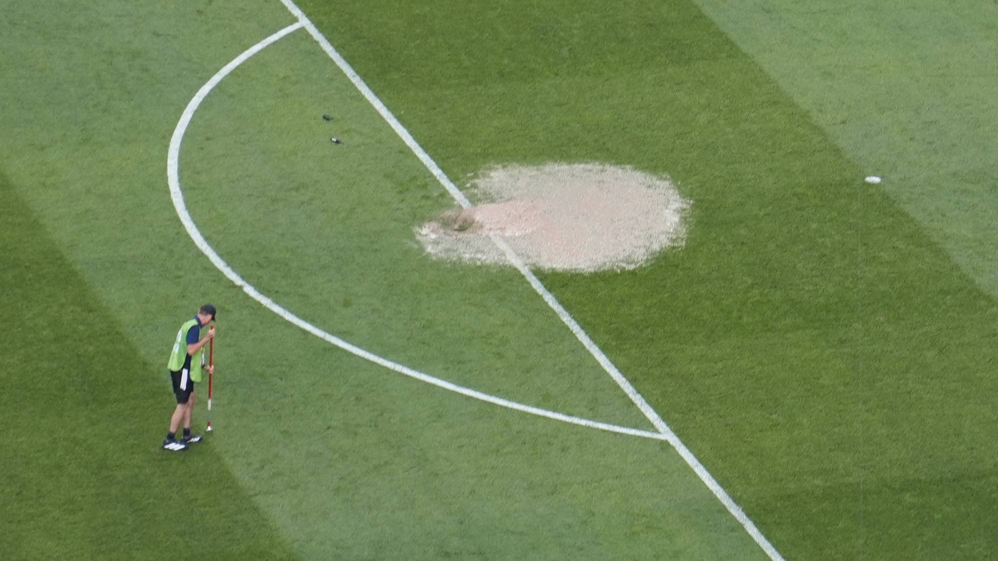 En fotballbane med flere personer. En står ved straffemerket mens en annen står ved straffefeltet. Ved straffefeltet er det en stor vanndam. I bakgrunnen er det et må l, publikum og reklameskilt. (Bildebeskrivelsen er laget av en KI-tjeneste)