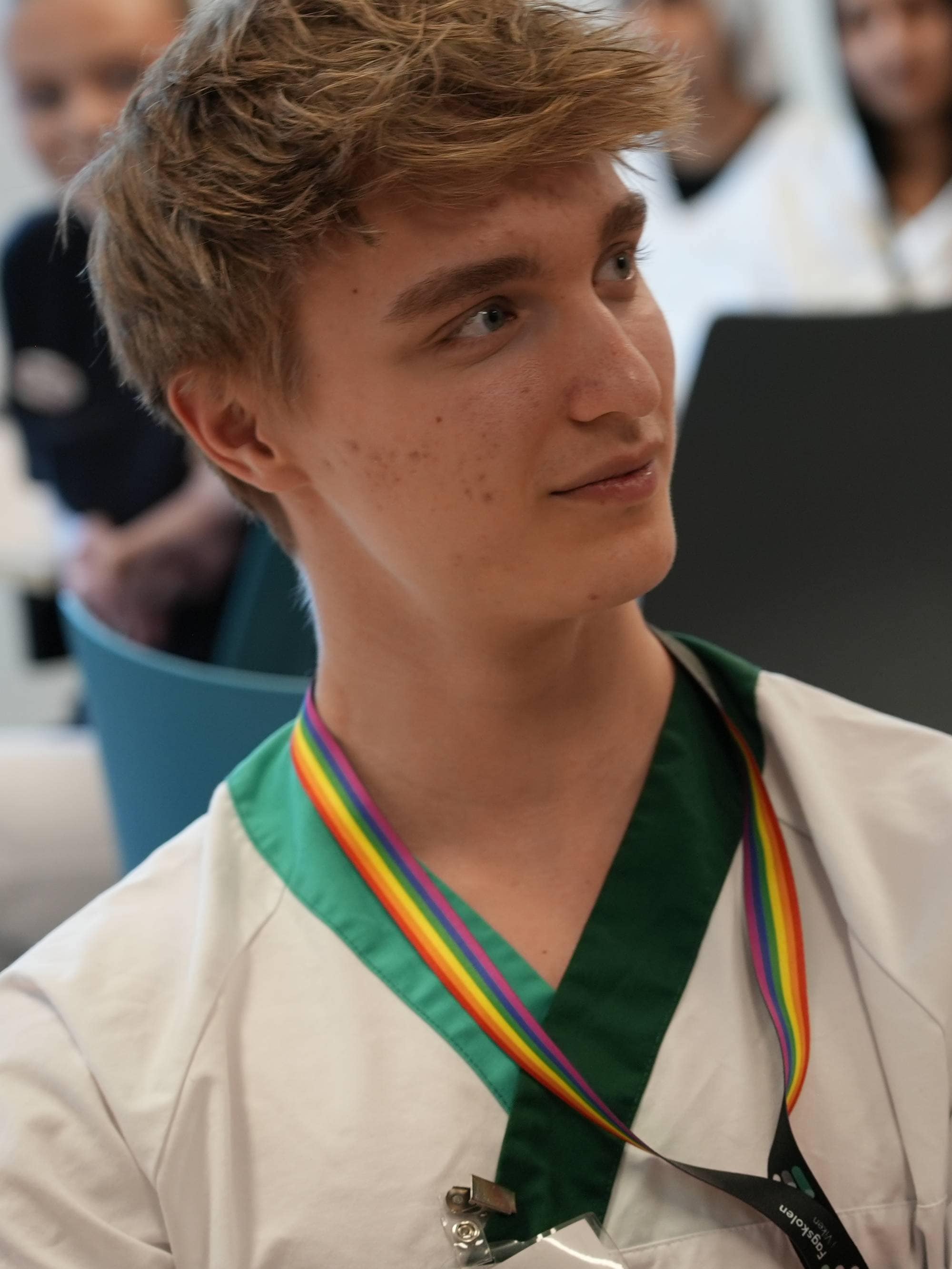 En ung mann med fregner og blondt hår sitter i et rom. Han har på seg en hvit skjorte med grønn krage og et regnbuefarget bånd rundt halsen. I bakgrunnen sitter flere personer, noen av dem i hvite klær. (Bildebeskrivelsen er laget av en KI-tjeneste)