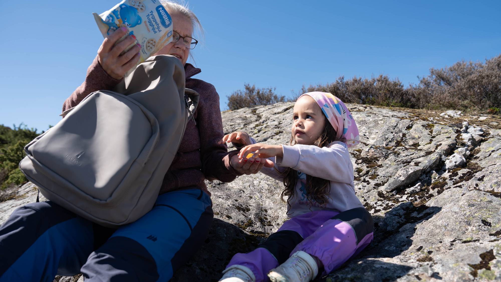 Ei kvinne gir ei jente ein matbit. Dei sit på ein stein i naturen. Kvinna har ein grå sekk på fanget. 