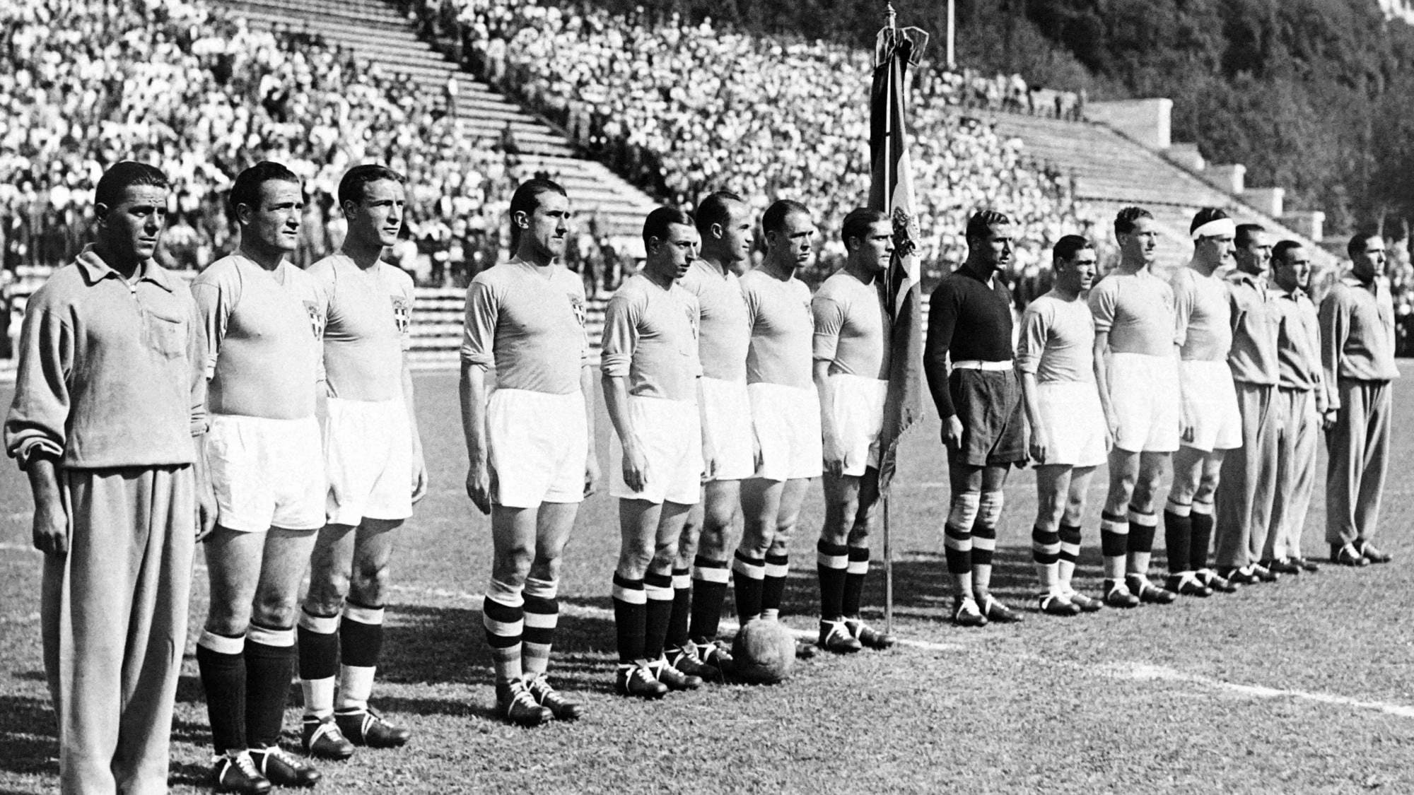 Et lag med fotballspillere står på en gressbane og ser fremover. En fane er plassert bak den første rekken av spillere, som står side om side. Tilskuere er synlige på tribunene i bakgrunnen. Bildet er i svart-hvitt. (Bildebeskrivelsen er laget av en KI-tjeneste)