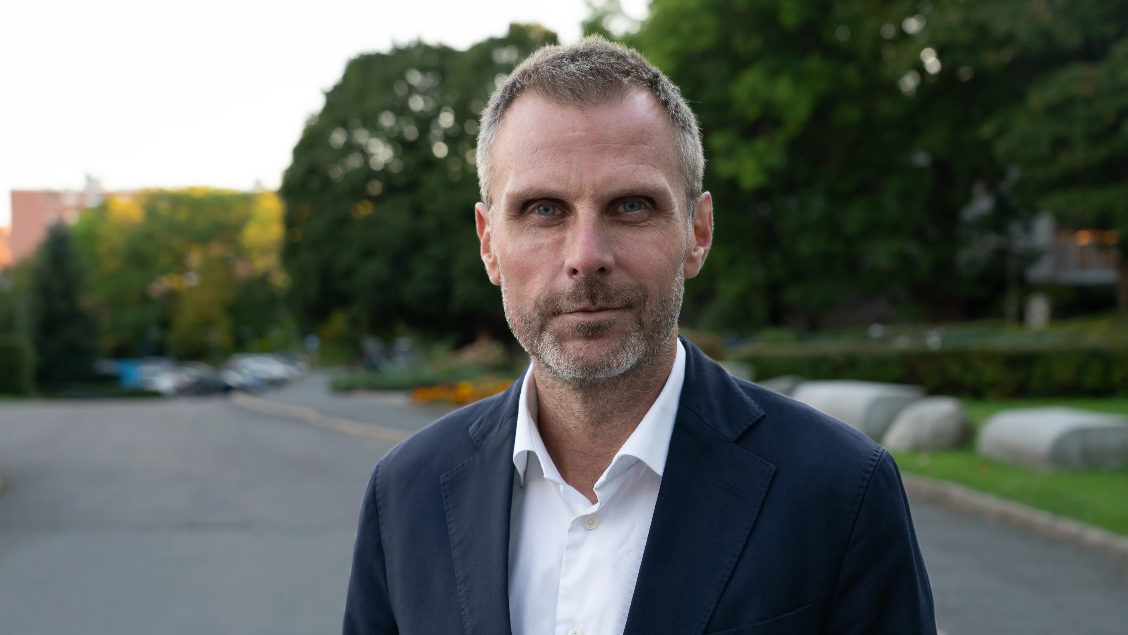 En mann i hvit skjorte og blå dressjakke ser rett i kamera med et lite smil om munnen.