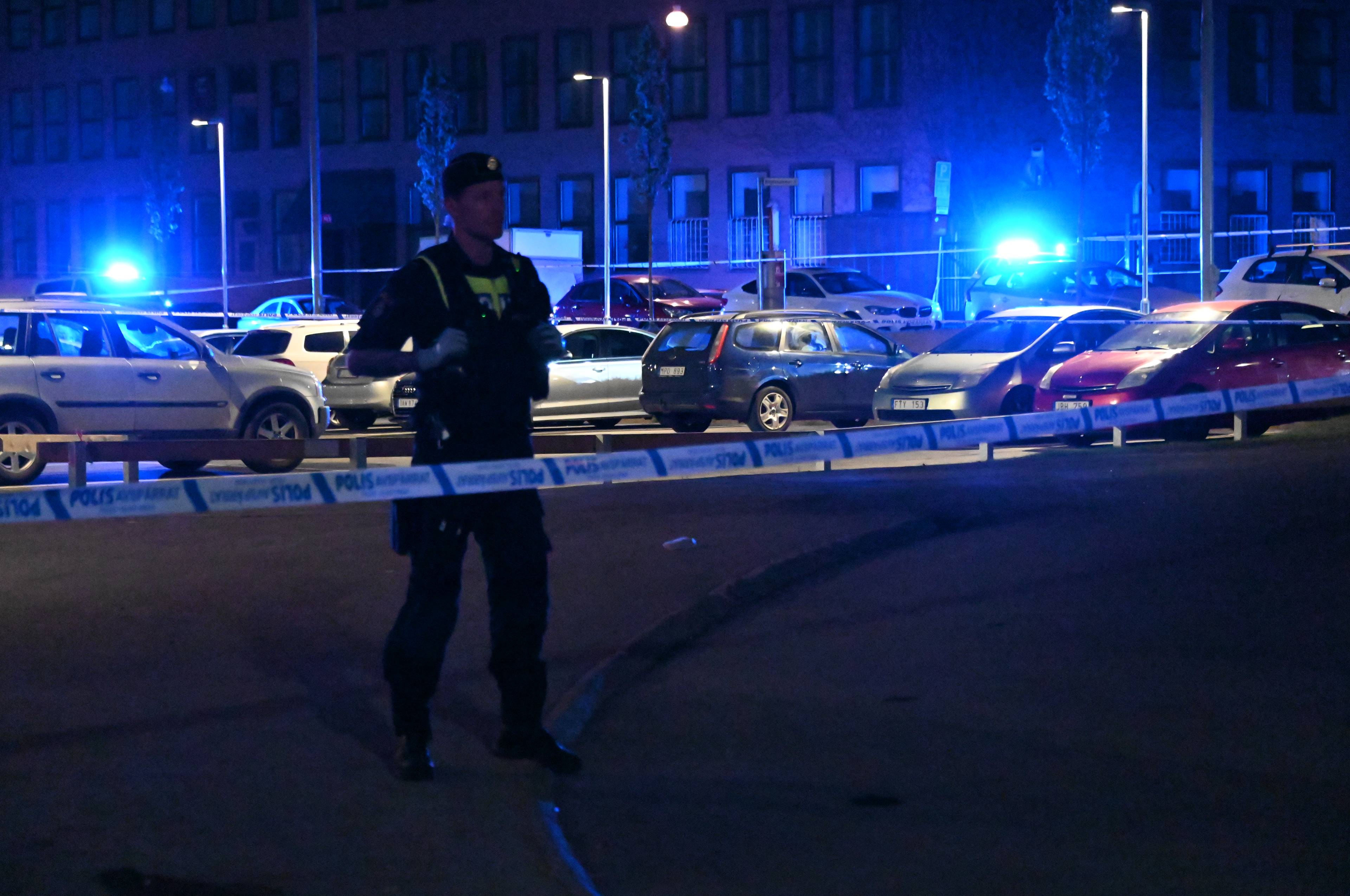 En politimann står ved et sperret område med politibånd. Flere biler er parkert bak sperringen, og et boligbygg med blått lys ses i bakgrunnen.