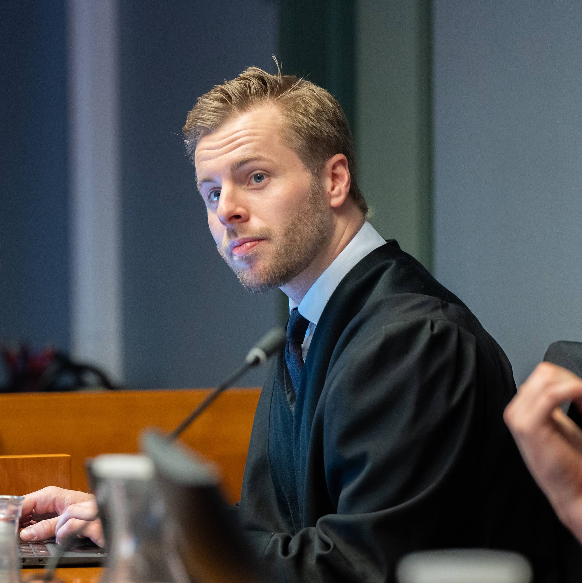 Advokat i domstolskappe sitter ved et bord og ser mot en person som snakker.