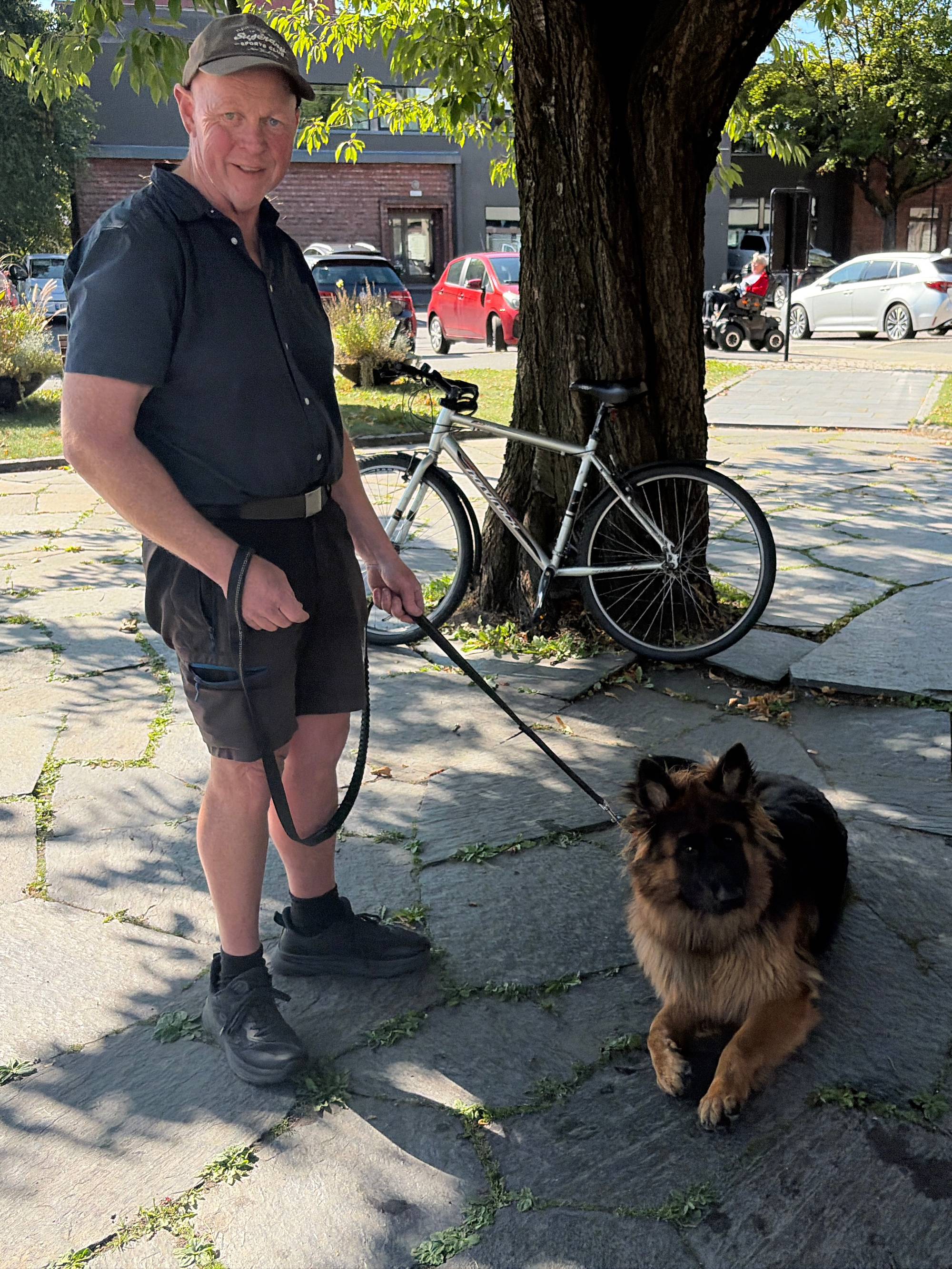 Mann med caps og hund i bånd står ved et tre på en steinlagt plass, med en sykkel bak seg. (Bildebeskrivelsen er laget av en KI-tjeneste)
