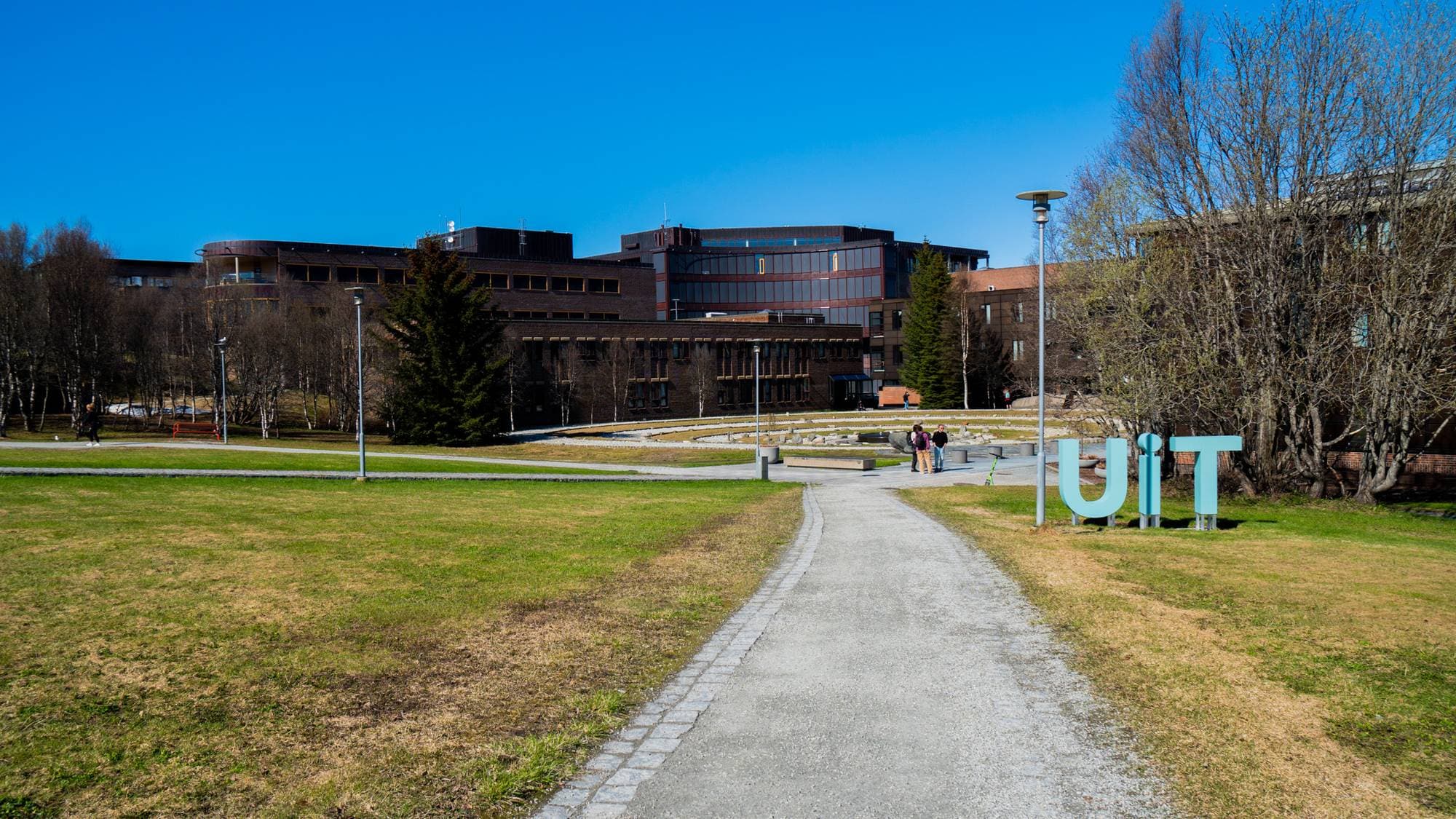 UiT Norges Arktiske Universitet campus Tromsø. Det er en grussti som går inn mot midten av bildet. På begge sidene av stien er det grønt og litt brunt gress. Det står UiT i store turkise bokstaver til høyre i bildet foran flere bygninger. Bygningene er i ulike størrelser og ulike bruntoner. 