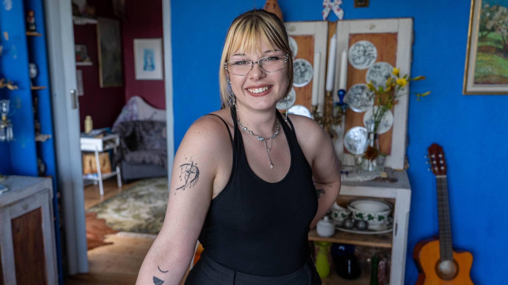 Kvinne med briller og tatoveringer på armen smiler i et rom med blå vegger. (Bildebeskrivelsen er laget av en KI-tjeneste)