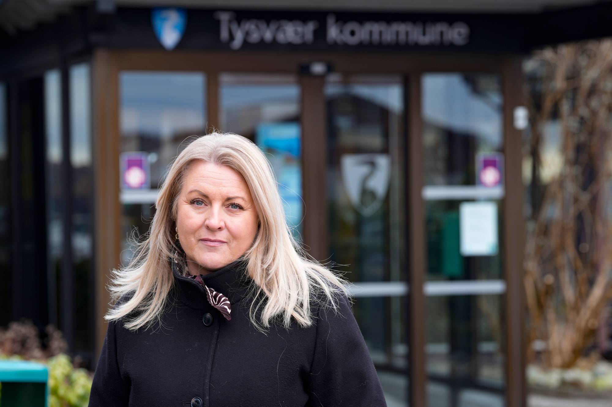Kvinne står foran Tysvær kommune-bygget. Hun har blondt hår og er kledd i en svart jakke. Bak henne skimtes inngangspartiet med glassdører og en tavle. (Bildebeskrivelsen er laget av en KI-tjeneste)
