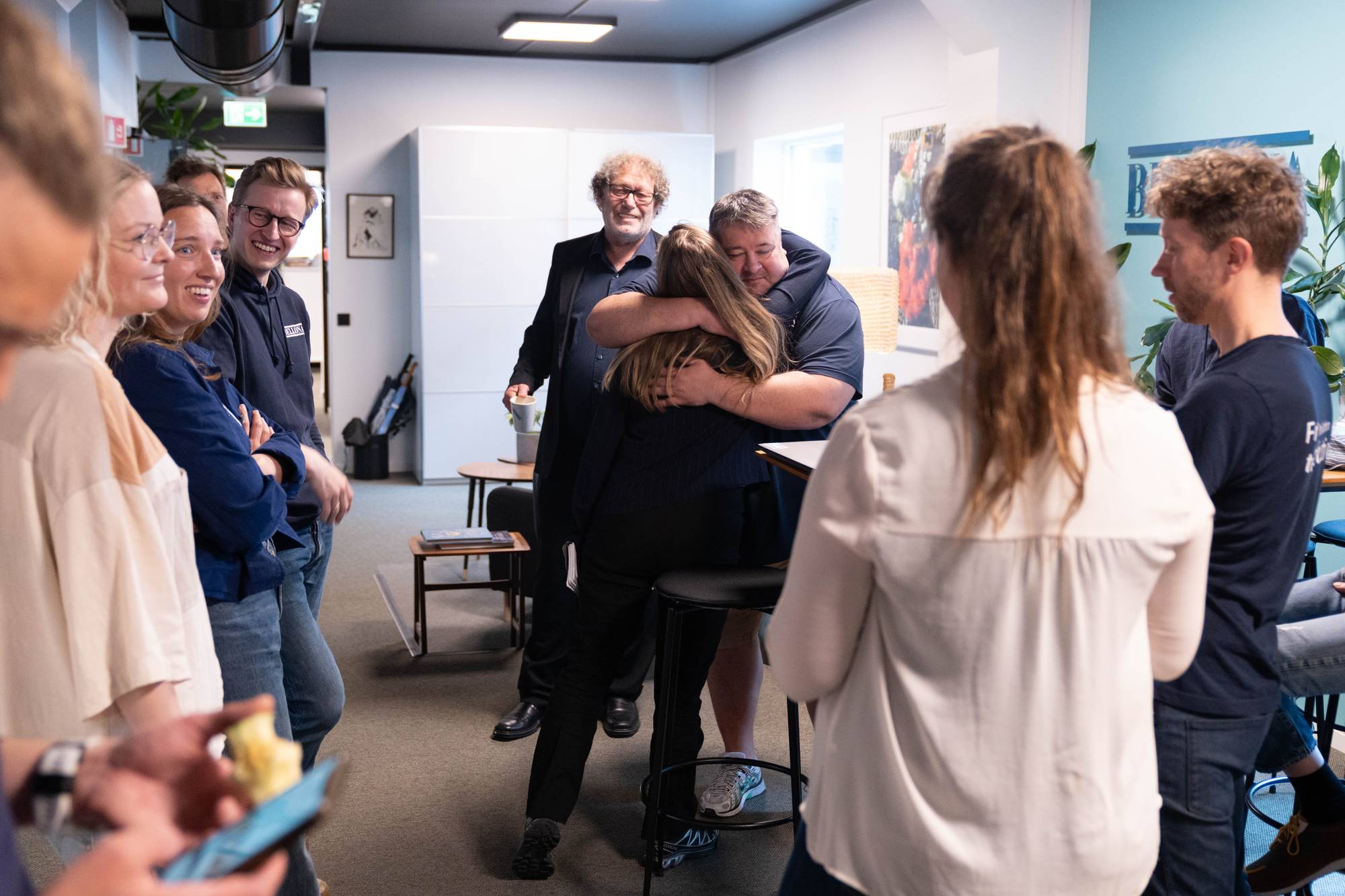 En mann i dress gir en kvinne en klem, mens en annen mann med krøllete hår ser på. Rundt dem står en gruppe mennesker og ser på. De befinner seg innendørs, muligens på et kontor. (Bildebeskrivelsen er laget av en KI-tjeneste)