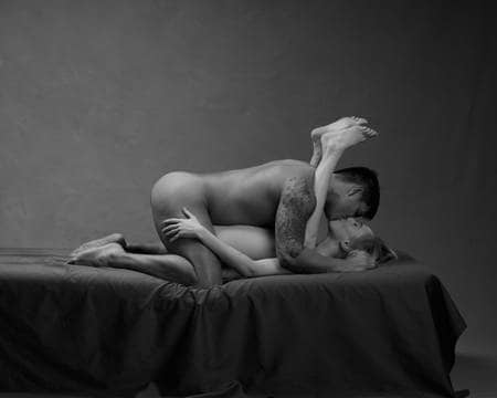 En naken dame ligger på ryggen ryggen med bena bretta opp rundt nakken til en naken mann som kysser henne