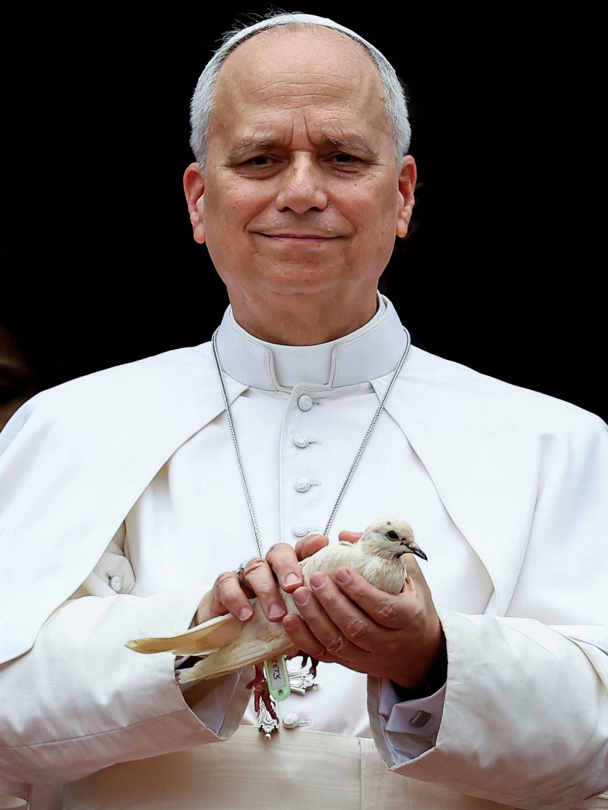 En mann i hvit geistlig drakt holder en hvit due i hendene. Han smiler mot kamera. Bildet er tatt utendørs foran en mørk bakgrunn. (Bildebeskrivelsen er laget av en KI-tjeneste)