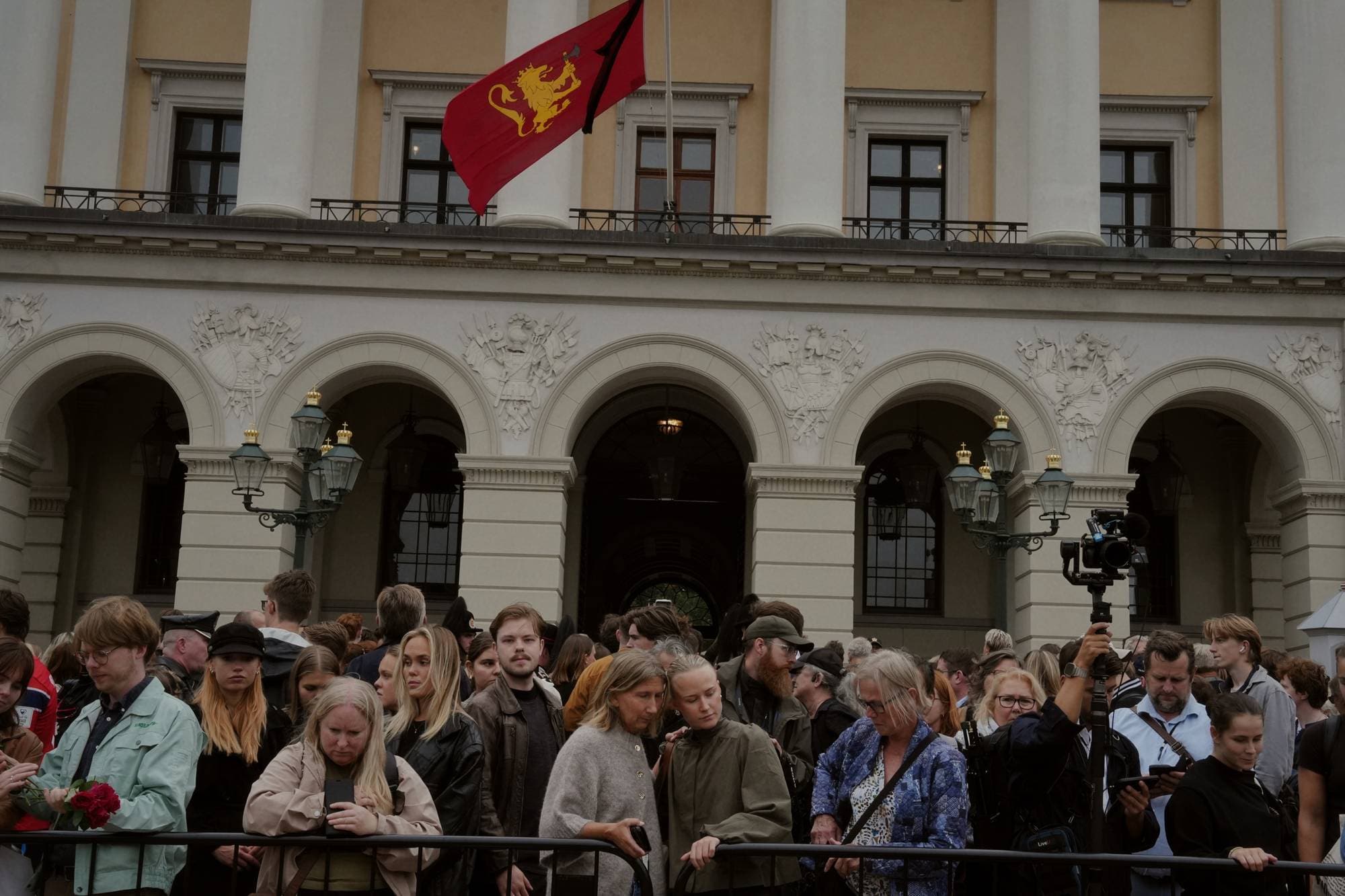 Mange mennesker står samlet utenfor Slottet. Noen holder rundt hverandre. I bakgrunnen sees kongeflagget med sørgeflor hengende fra slottsbalkongen.