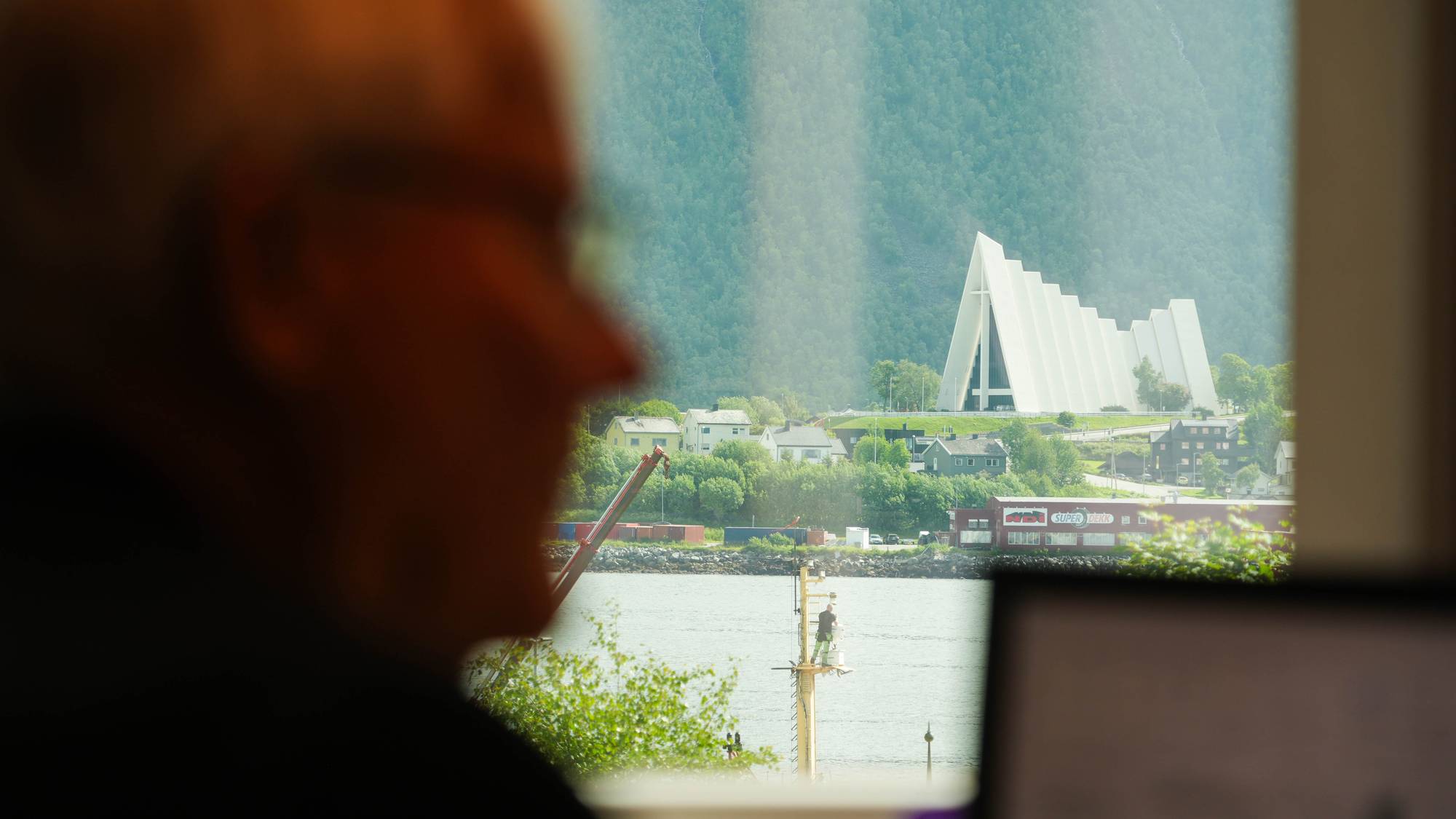 Utsikt mot Ishavskatedralen i Tromsø fra et vindu med en uklar person i forgrunnen. (Bildebeskrivelsen er laget av en KI-tjeneste)
