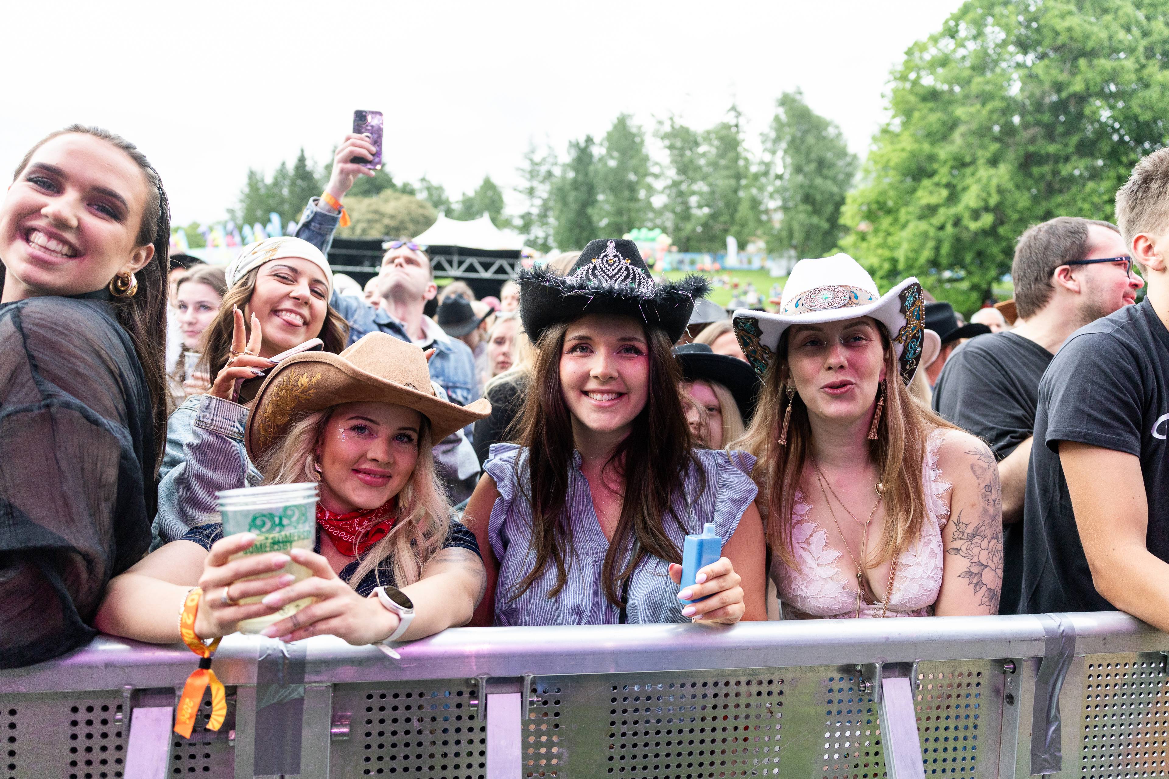 Tre kvinner, kledd i cowboystil, smiler mot kamera på en utendørs konsert. De står foran et publikum bak en scenebarriere. (Bildebeskrivelsen er laget av en KI-tjeneste)