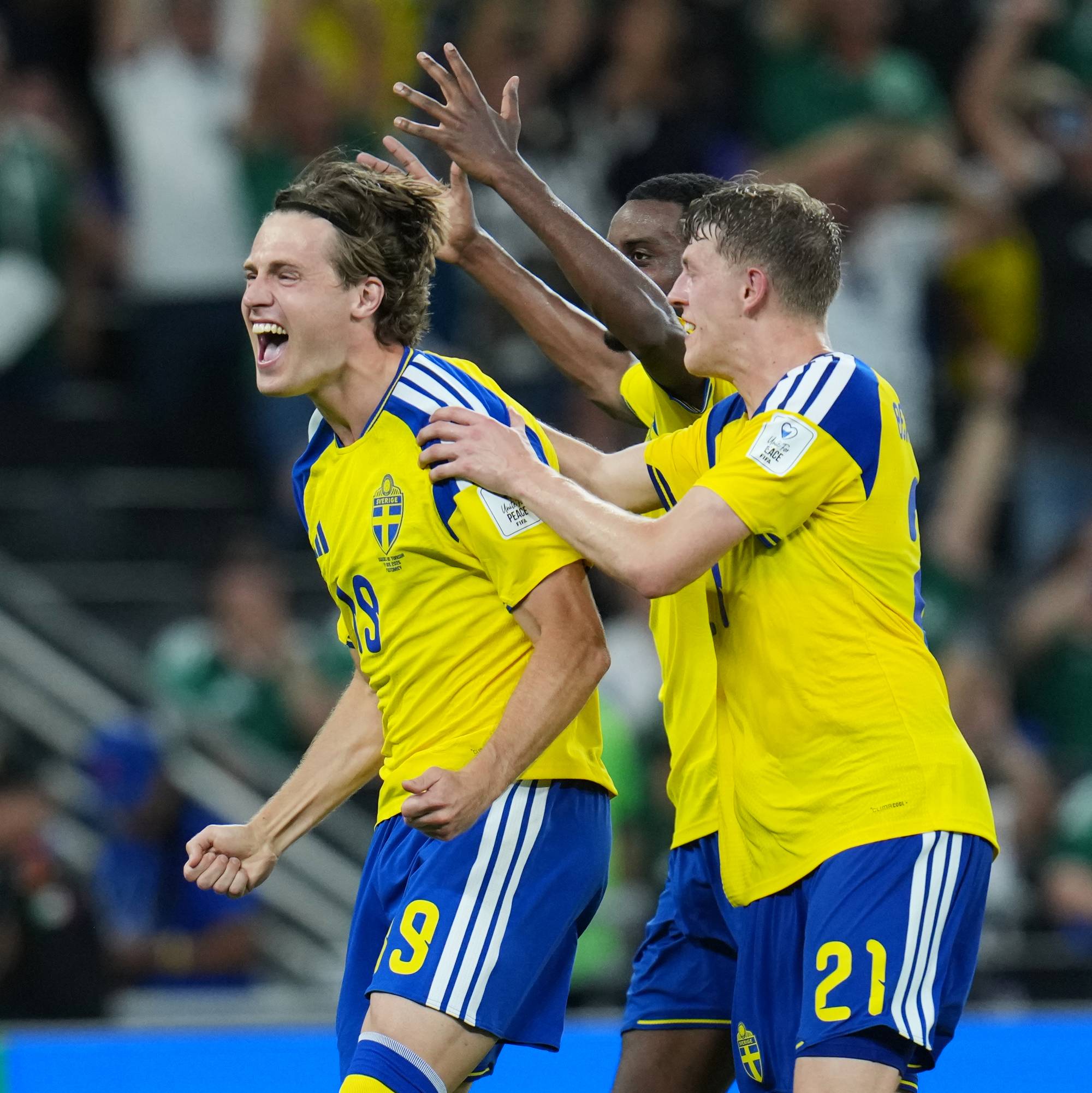 Tre svenske fotballspillere feirer et mål på en bane. Spilleren i forgrunnen, med nummer 19 på drakten, jubler med armene i været. En annen spiller, med nummer 21, klapper ham på skulderen. I bakgrunnen sees tilskuere på tribunen. (Bildebeskrivelsen er laget av en KI-tjeneste)