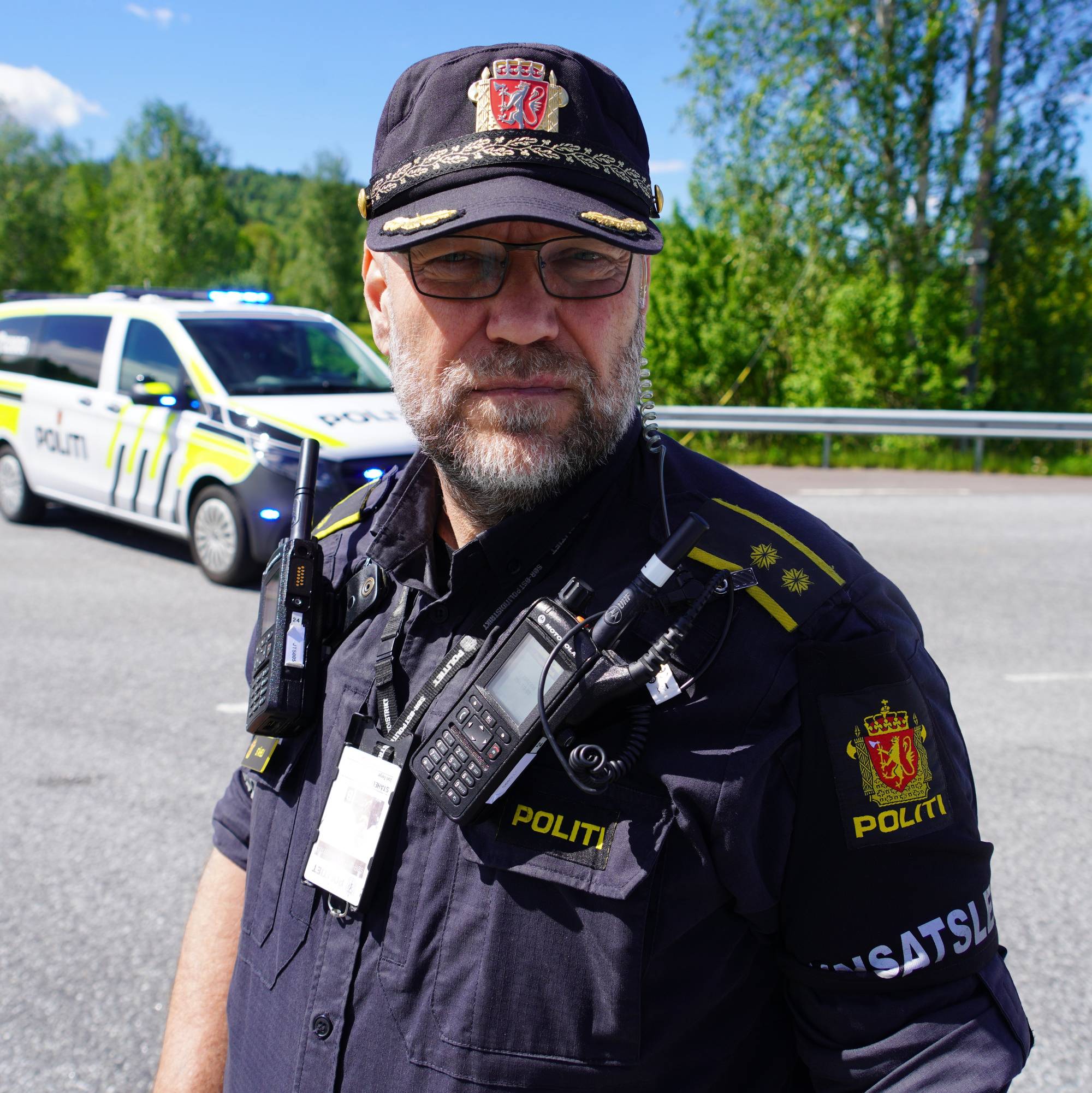 En politimann står utendørs med en politibil bak seg