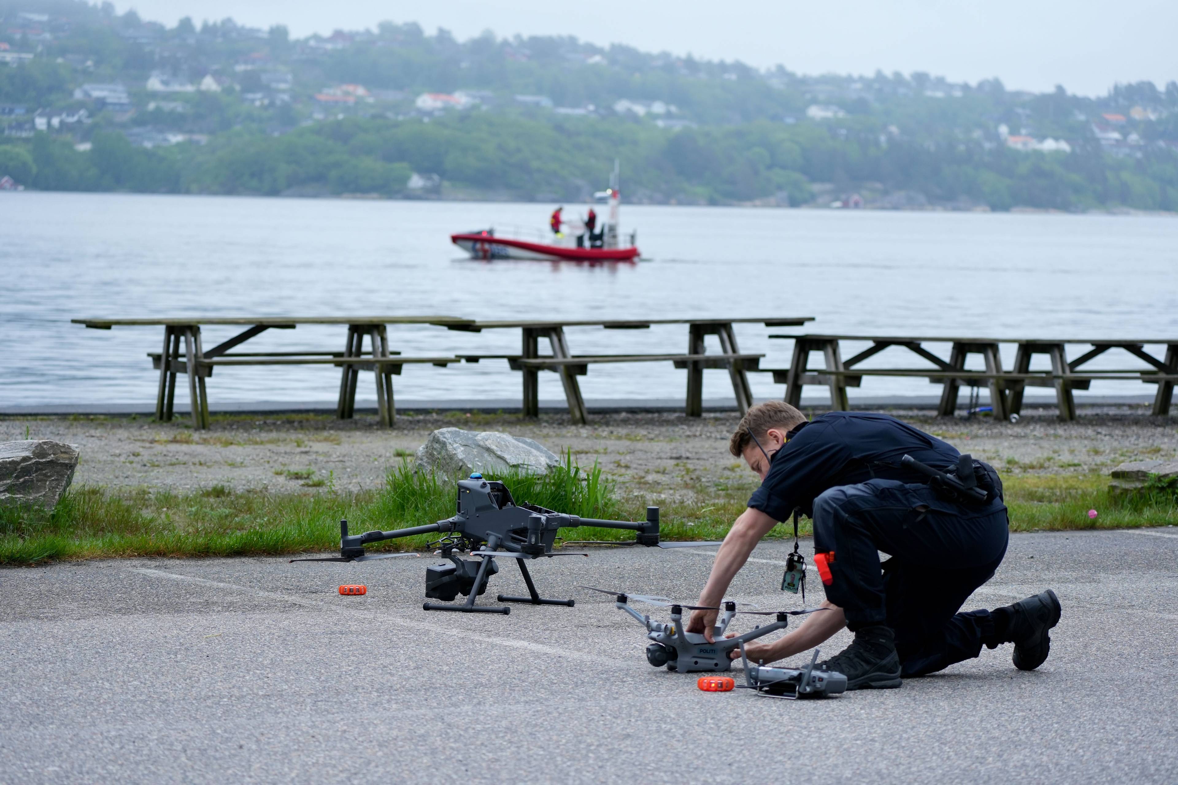 Politi i mørk uniform monterer en drone. Han er på et jorde med flere droner og redningsutstyr. I bakgrunnen er et vann med en båt. (Bildebeskrivelsen er laget av en KI-tjeneste)