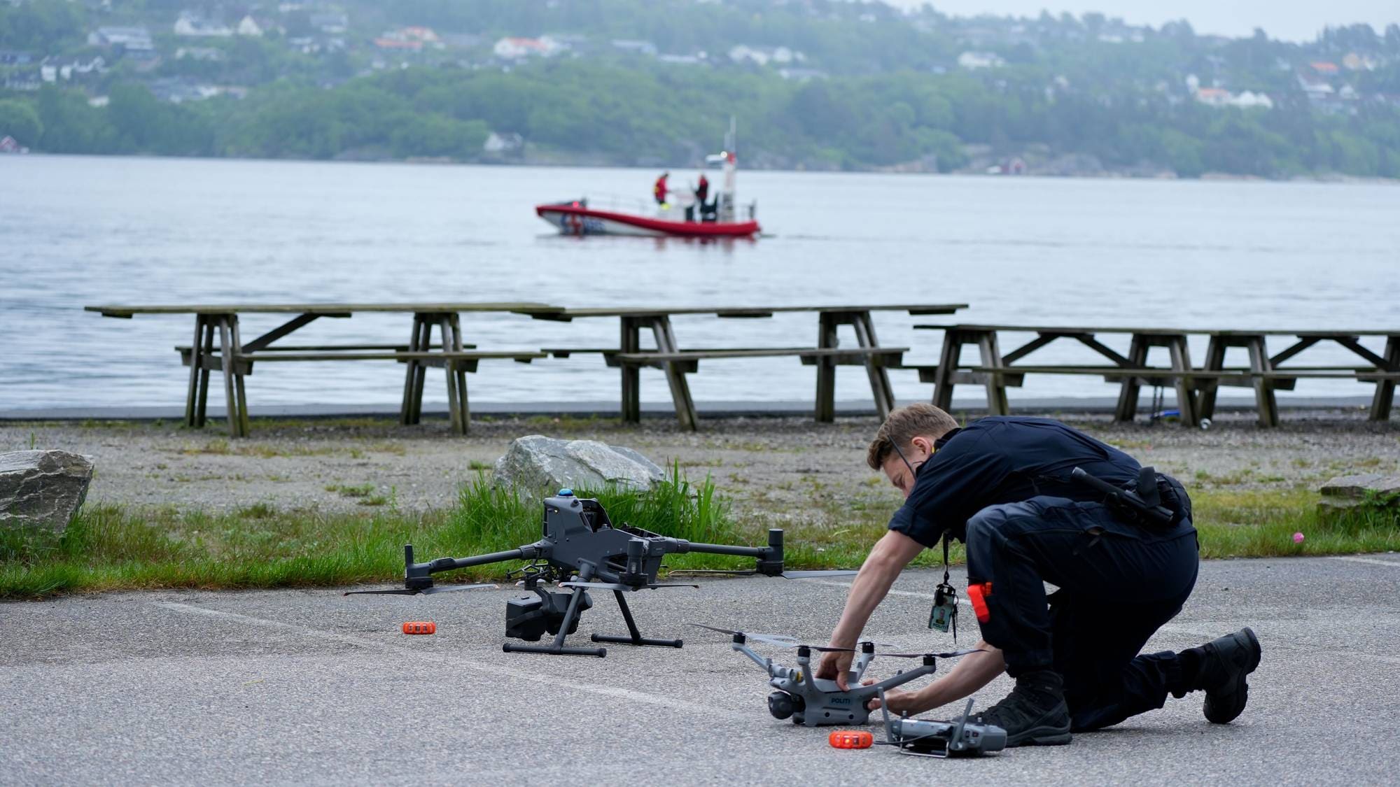 Politi i mørk uniform monterer en drone. Han er på et jorde med flere droner og redningsutstyr. I bakgrunnen er et vann med en båt. (Bildebeskrivelsen er laget av en KI-tjeneste)