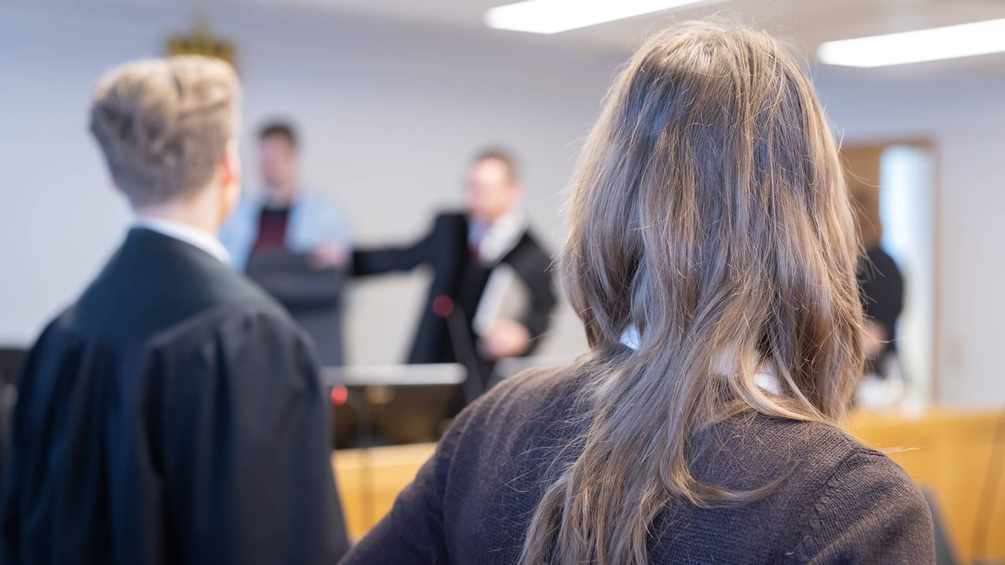En kvinne er fornærmet i en straffesak. Hun har reist seg i det dommerne kommer inn i rettssalen. Vi ser hennes bakhode og deler av ryggen, i tillegg til hennes bistandsadvokat. I bakgrunnen ser vi dommerne komme inn i rettssalen
