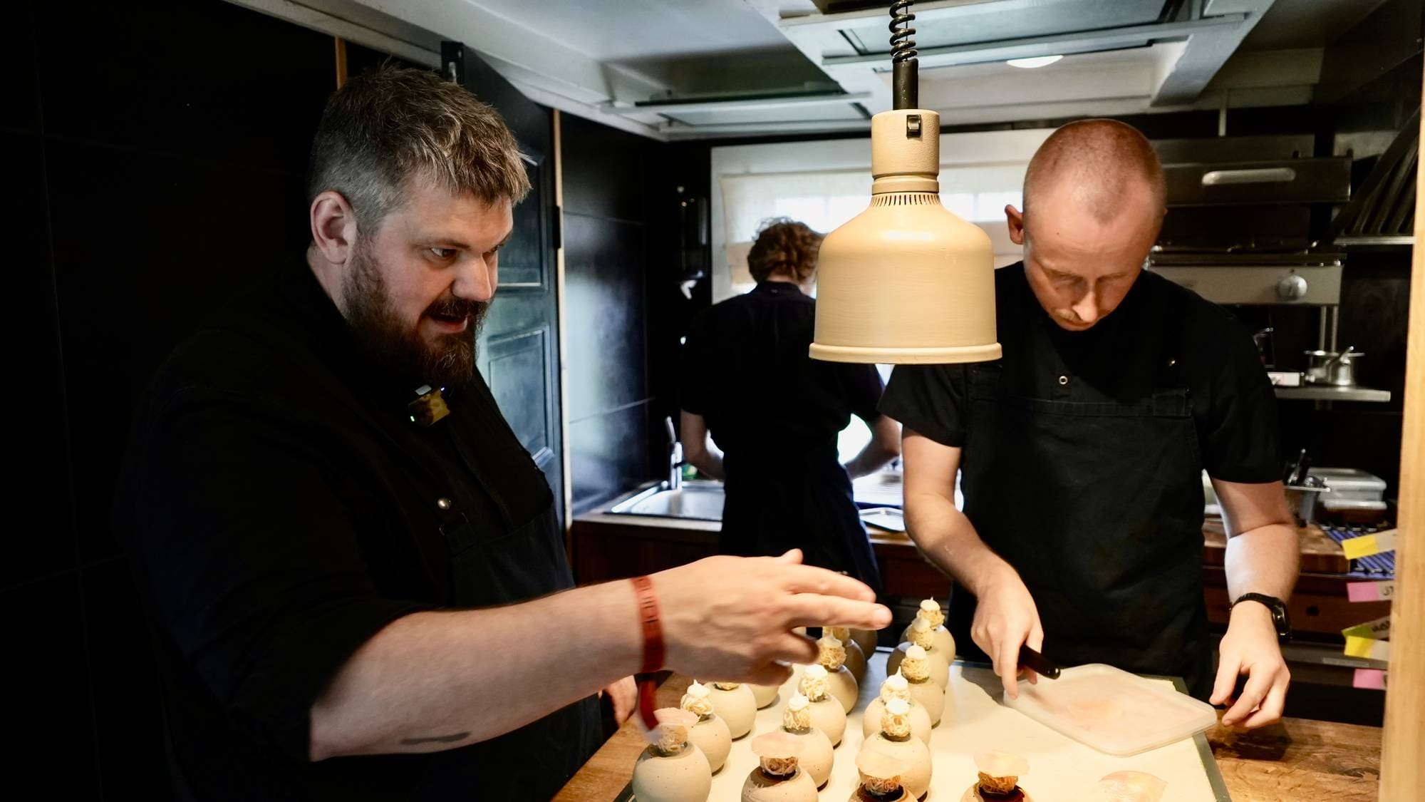 To kokker i svarte kokkejakkker jobber på et kjøkken. En mann til venstre peker på ferdiglagde desserter på et bord. En mann til høyre skjærer mat på et skjærebrett. (Bildebeskrivelsen er laget av en KI-tjeneste)