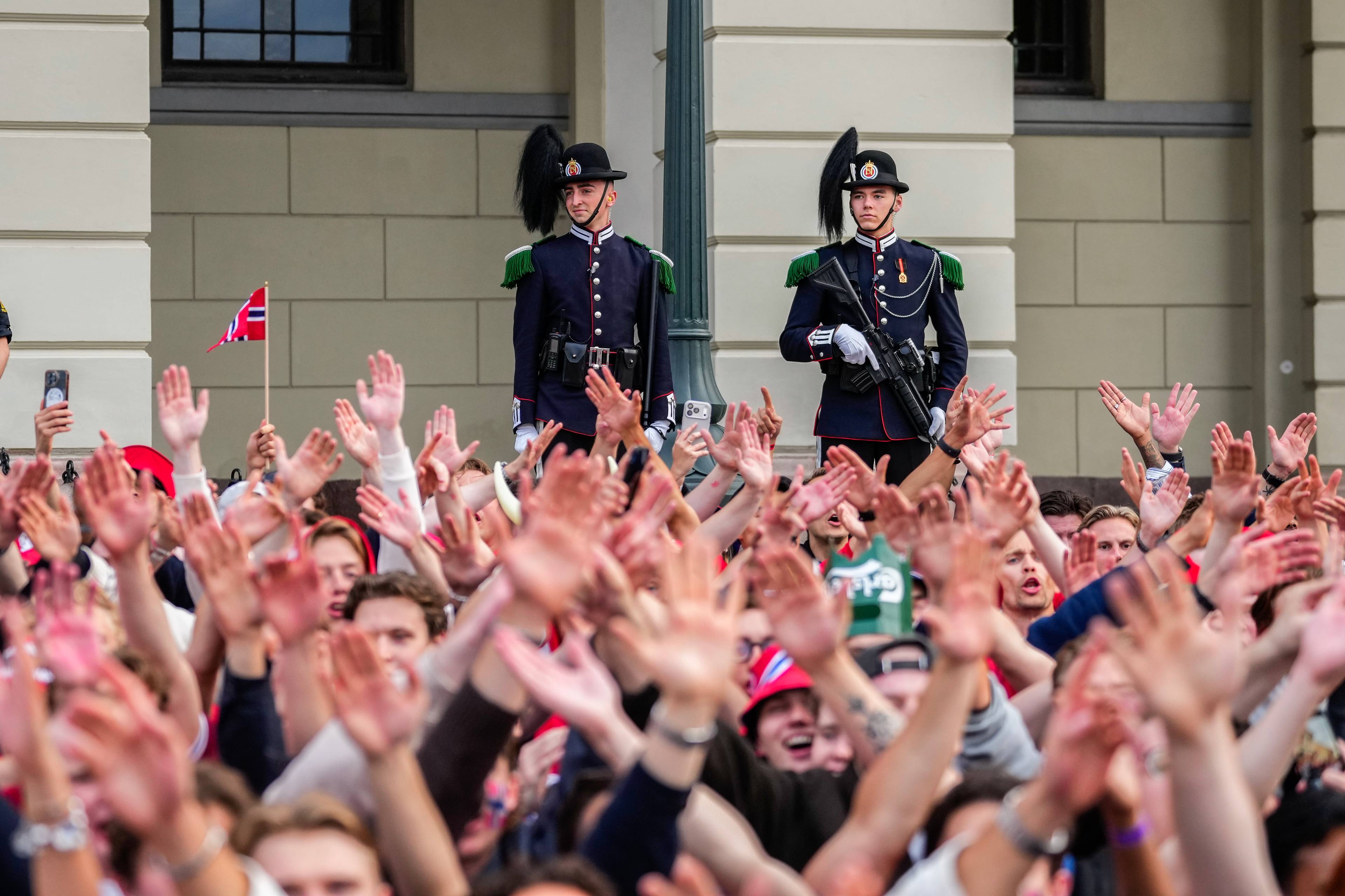 To vakter i uniform står bak en folkemengde. Folk har hendene løftet og det vaies med norske flagg. Bakgrunnen er en bygning.