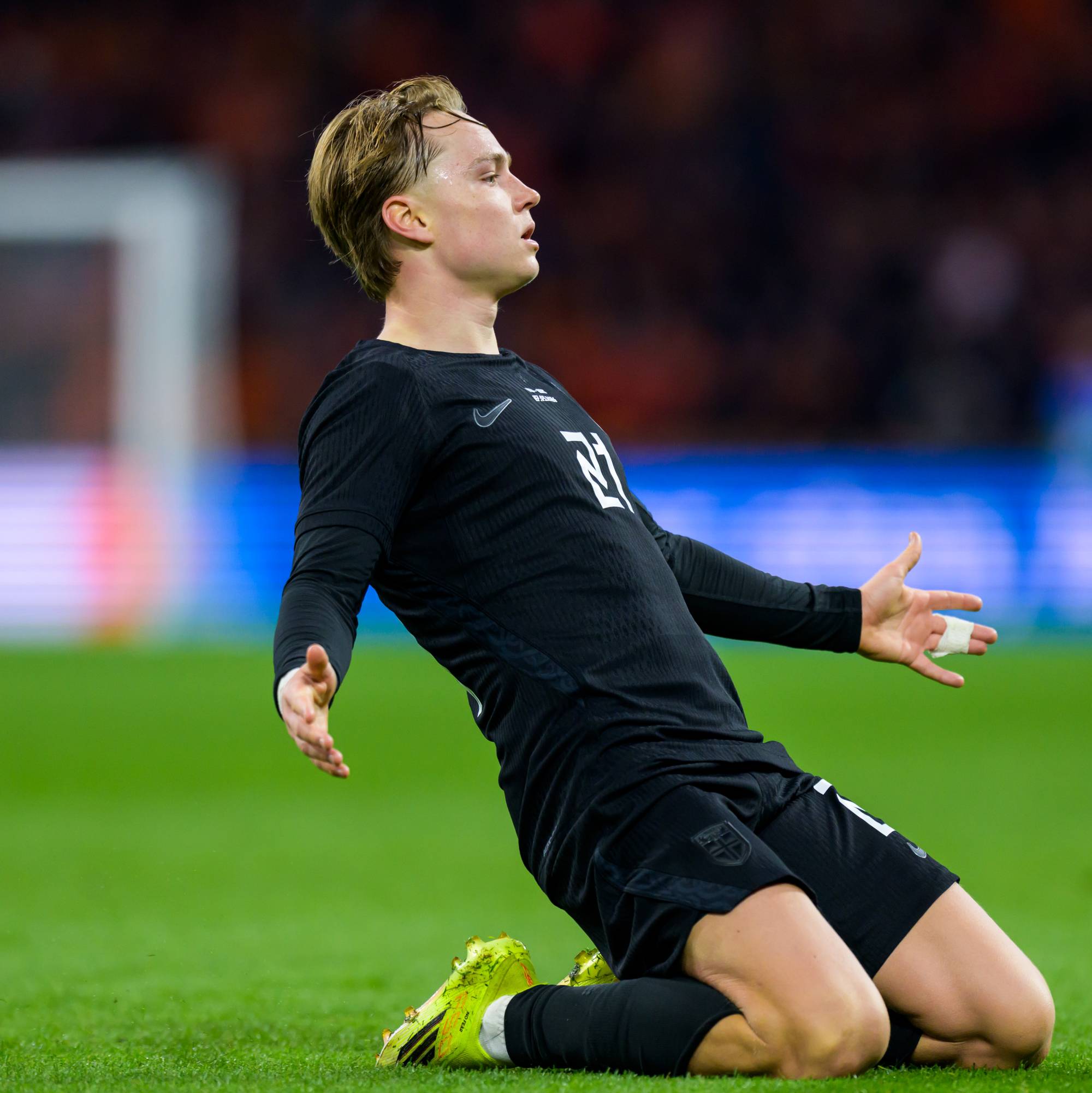 En fotballspiller i svart drakt kneler på en gressbane. Spilleren har blondt hår og ser ut til å være i bevegelse. Bak spilleren er det et utydelig publikum og et mål. (Bildebeskrivelsen er laget av en KI-tjeneste)