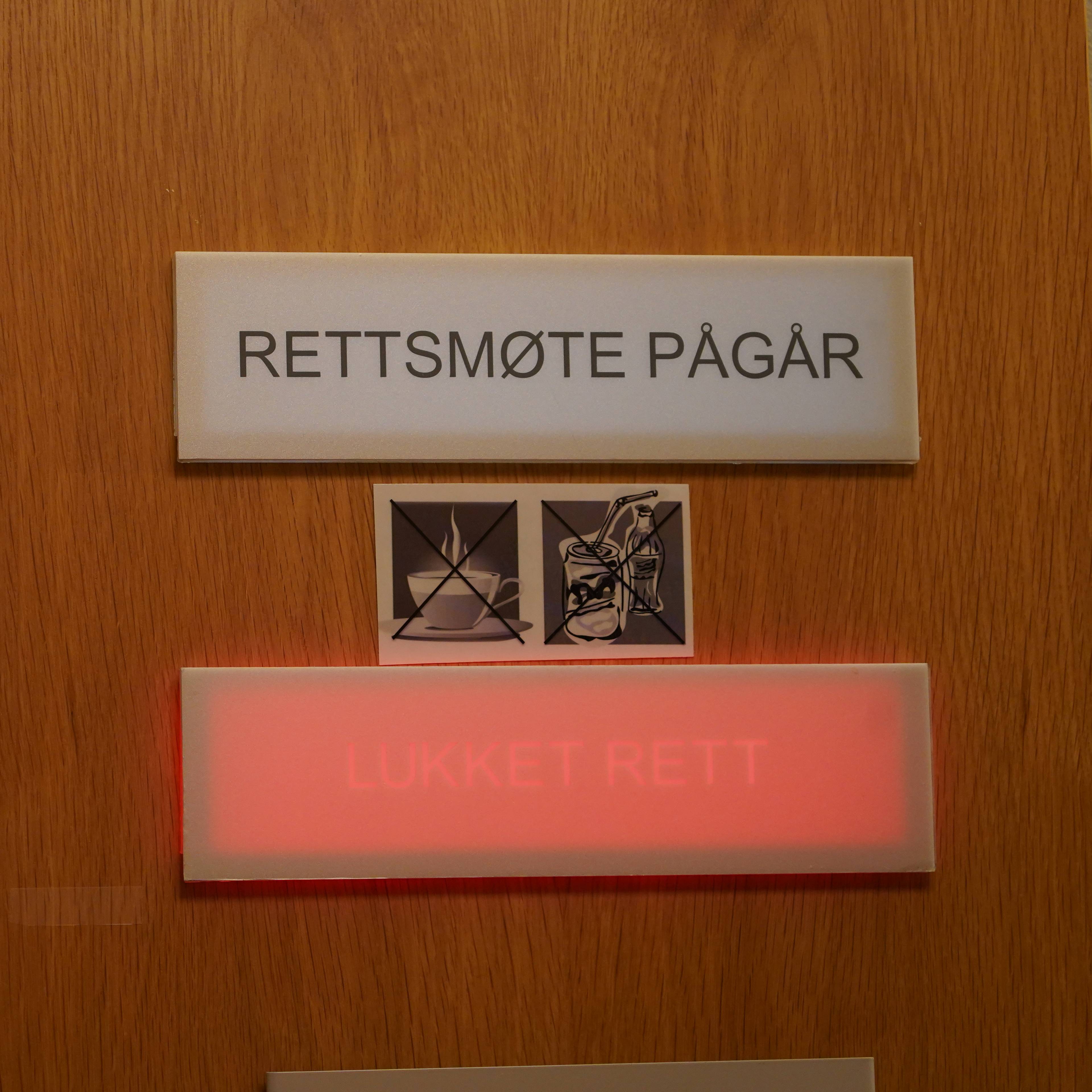 En dørskilt med teksten "RETTSMØTE PÅGÅR" er montert på en tredør. Under skiltet er det to små symboler. Ett symbol viser en kopp kaffe med en kryss over. Det andre symbolet viser en boks med sugerør og en flaske brus med en kryss over. Et rødt lysskilt under disse symbolene viser teksten "LUKKET RETT". (Bildebeskrivelsen er laget av en KI-tjeneste)
