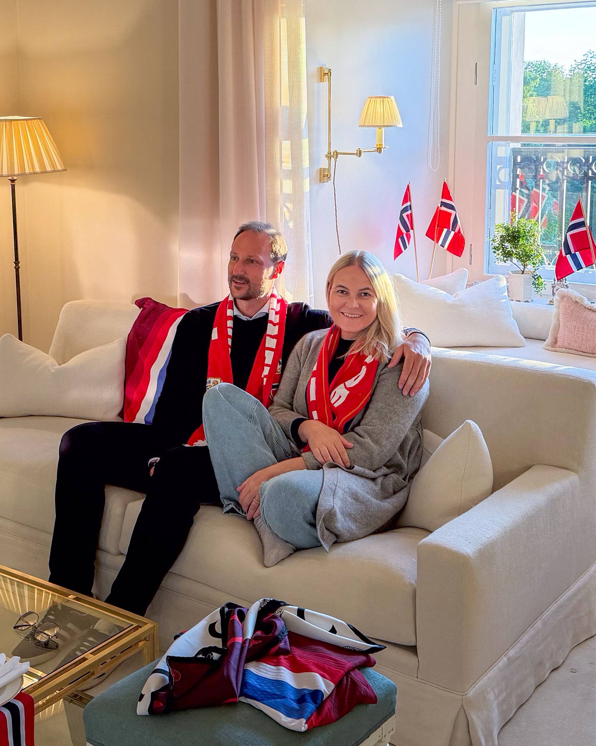 Kronprins Haakon og kronprinsesse Mette-Marit sitter i en sofa med norske flagg i bakgrunnen. (Bildebeskrivelsen er laget av en KI-tjeneste)