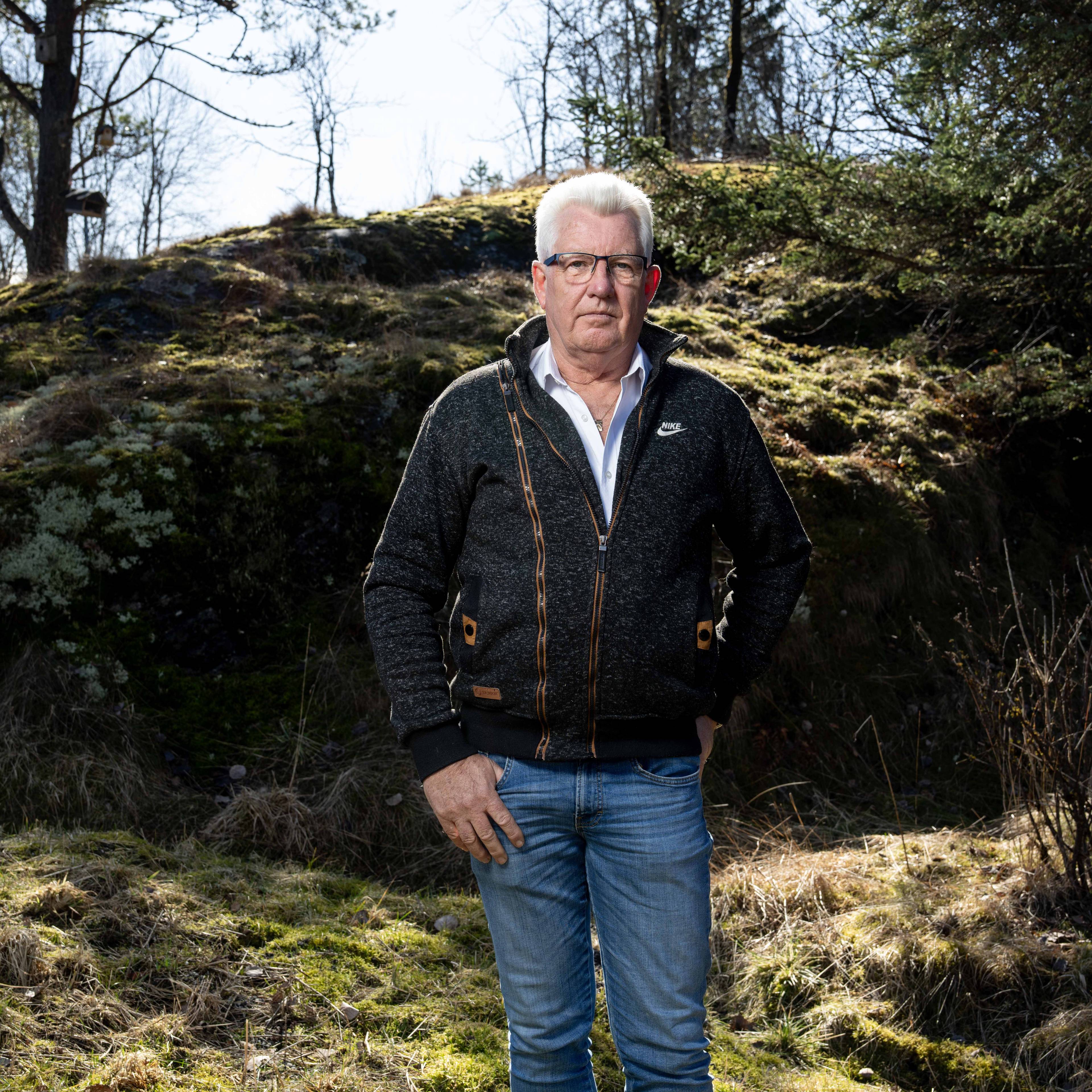 Erik Arntzen har på seg flis og hvit skjorte og står ute i skogen. Bak ham er en liten holme med trær. Det er vår og svakt sollys.