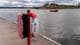Foto: Christian Nygaard-Monsen / NRK Livbøye og redningsutstyr står på en brygge ved vannet. I bakgrunnen er det røde hytter på en øy. En liten rød båt med en person ombord kjører forbi. (Bildebeskrivelsen er laget av en KI-tjeneste)