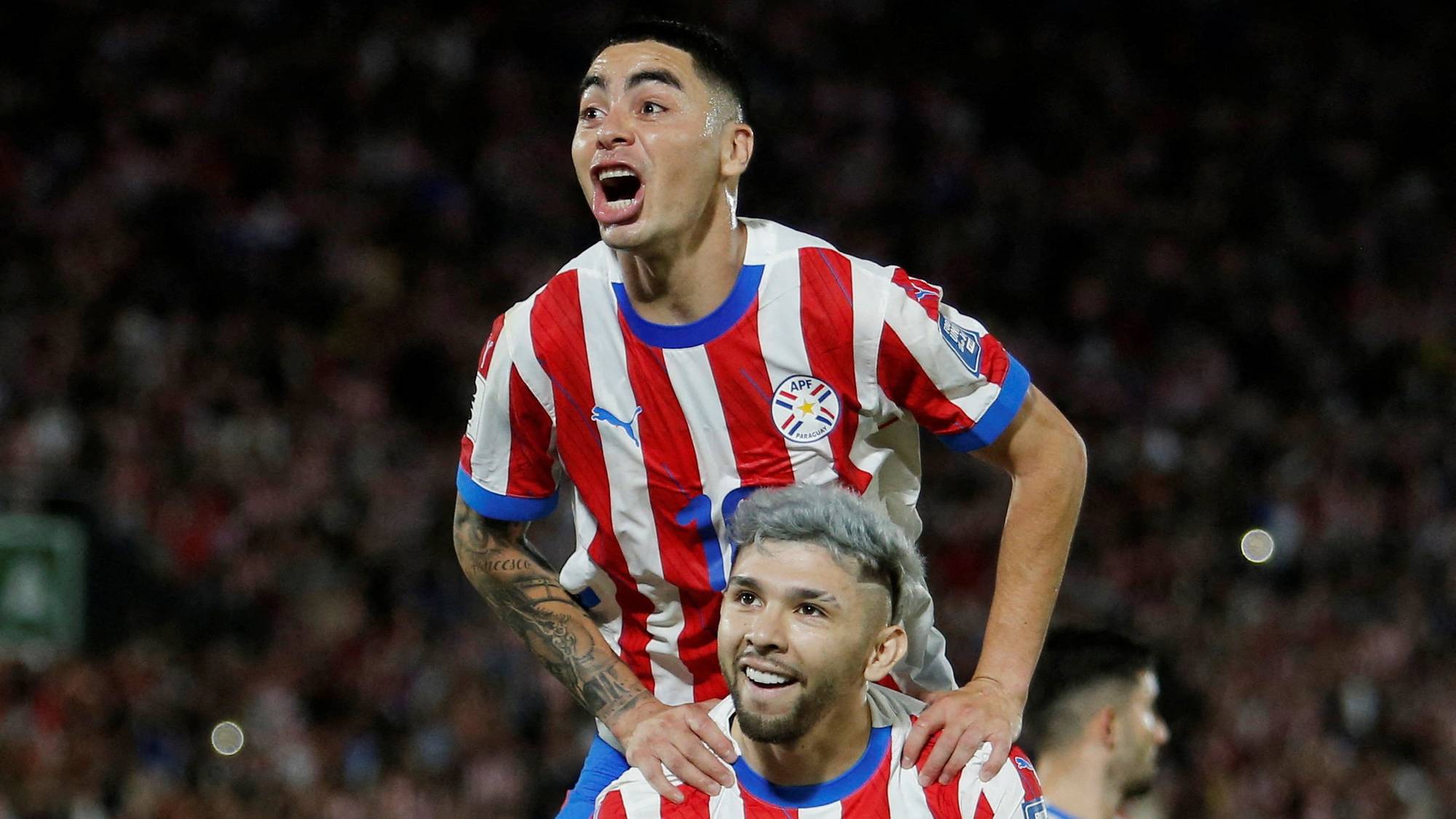 To fotballspillere i Paraguay-drakt feirer. En spiller er på skuldrene til en annen. Bakgrunnen viser en tribunefylt stadion. (Bildebeskrivelsen er laget av en KI-tjeneste)