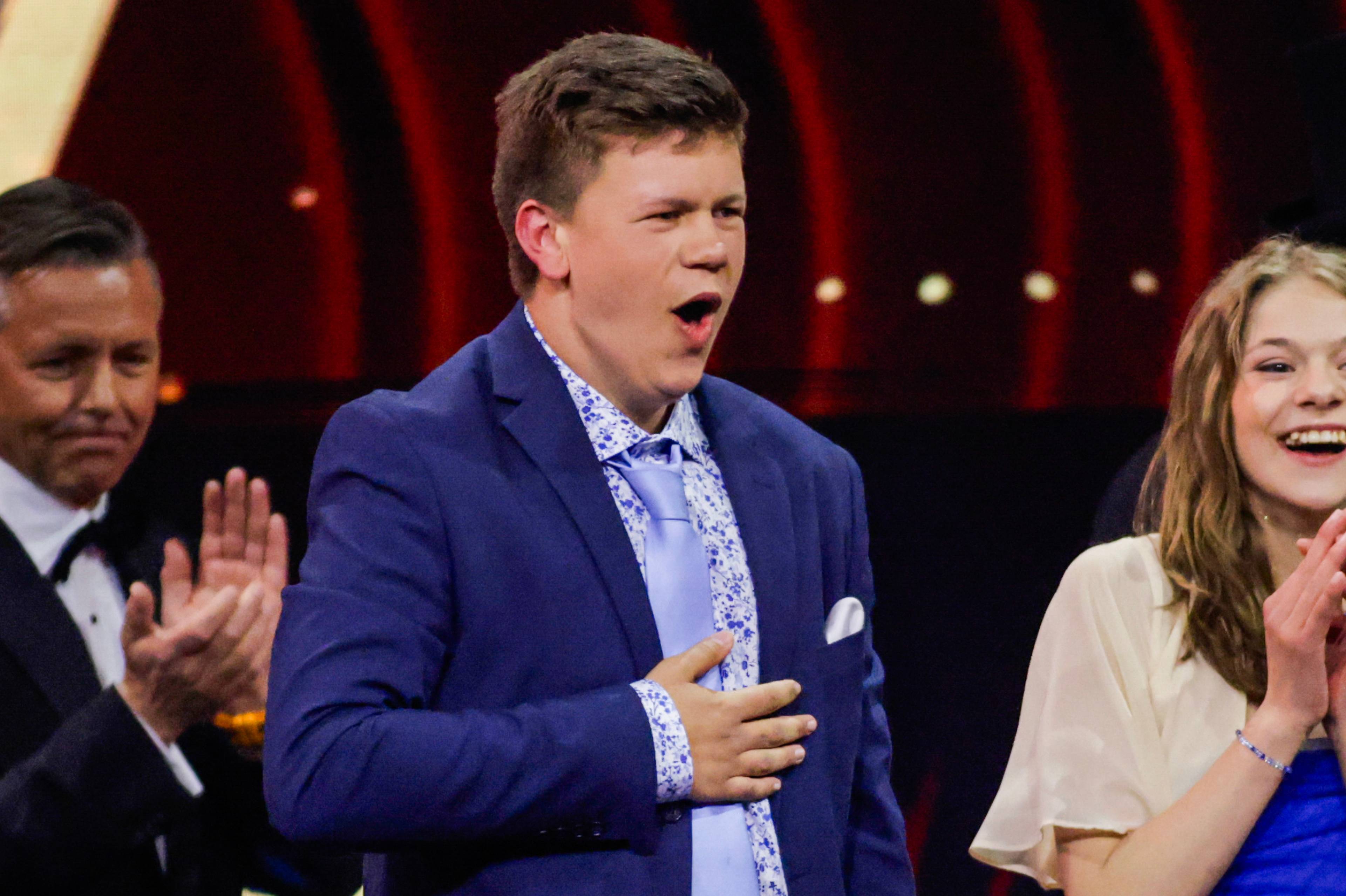 En ung mann i blå dress holder hånden på brystet og ser til høyre. Bak ham står en mann med sløyfe og en ung kvinne som klapper. Scenen har en mørk bakgrunn med røde lys. (Bildebeskrivelsen er laget av en KI-tjeneste)