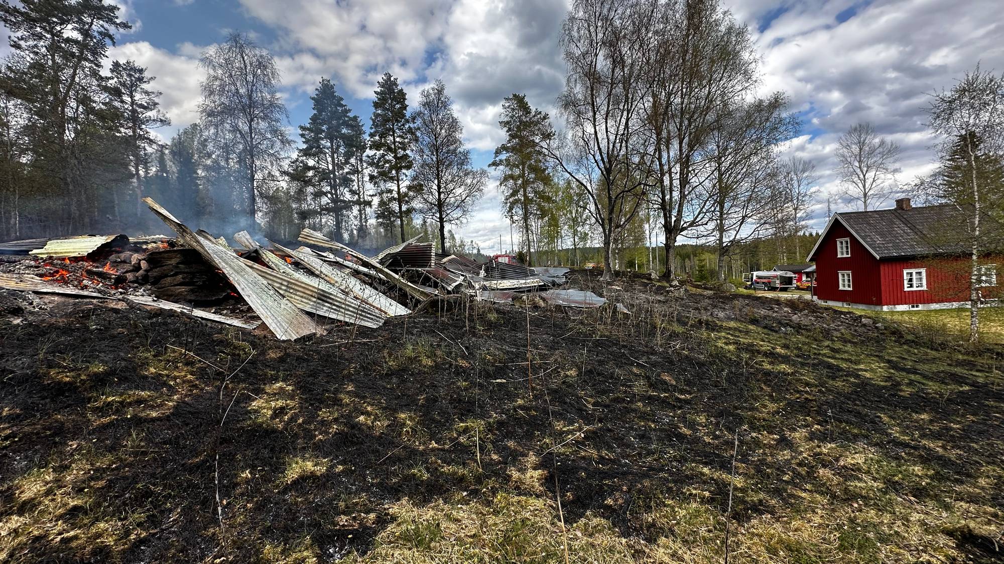 Rester av en bygning etter brann. I bakgrunnen ligger en rød bygning og brannbiler. (Bildebeskrivelsen er laget av en KI-tjeneste)