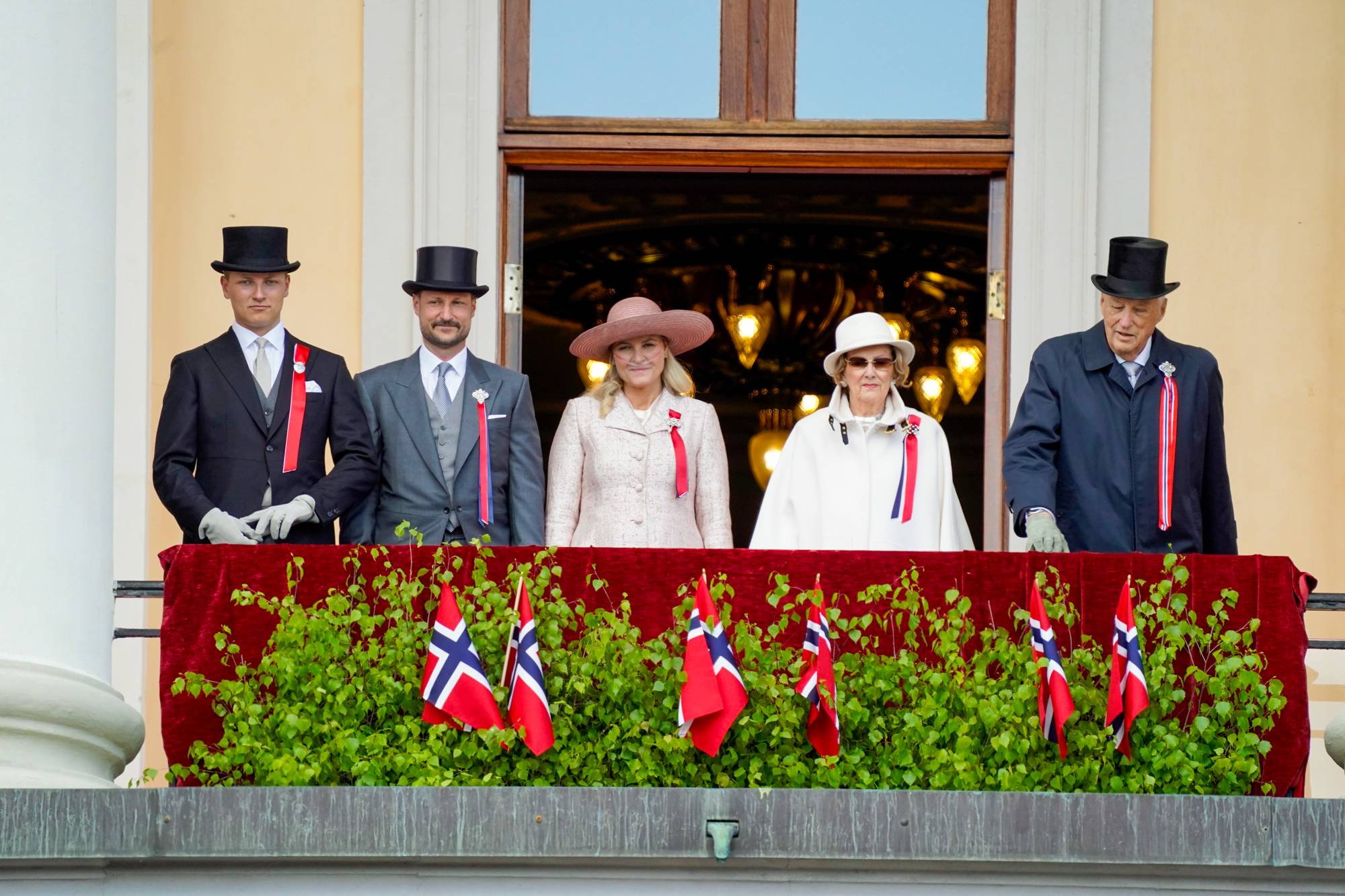 Kongefamilien på balkongen, mennene med flosshatt, mørke dresser og kronprinsessen og dronningen i hvite kåper og hatter.