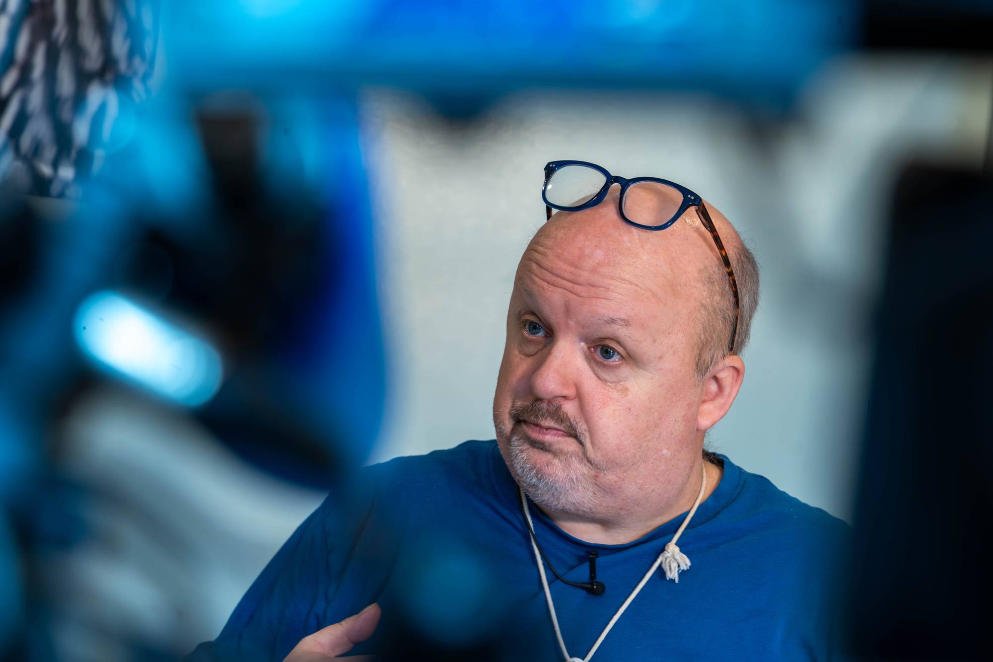 En mann med blå briller på hodet er avbildet i en blå genser. Mannen ser til venstre for kameraet med en nølende mine. Bildet er tatt tett på mannen, med blå og hvite farger i bakgrunnen.