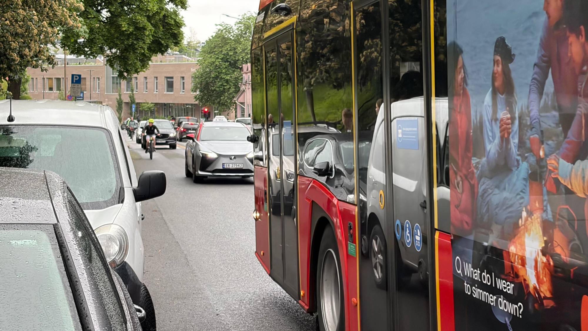 Trafikkork i byen. En person sykler forbi en buss med reklame. En bil med en Lexus-logo er synlig i trafikken. Det er regnvær, og veien er våt. (Bildebeskrivelsen er laget av en KI-tjeneste)