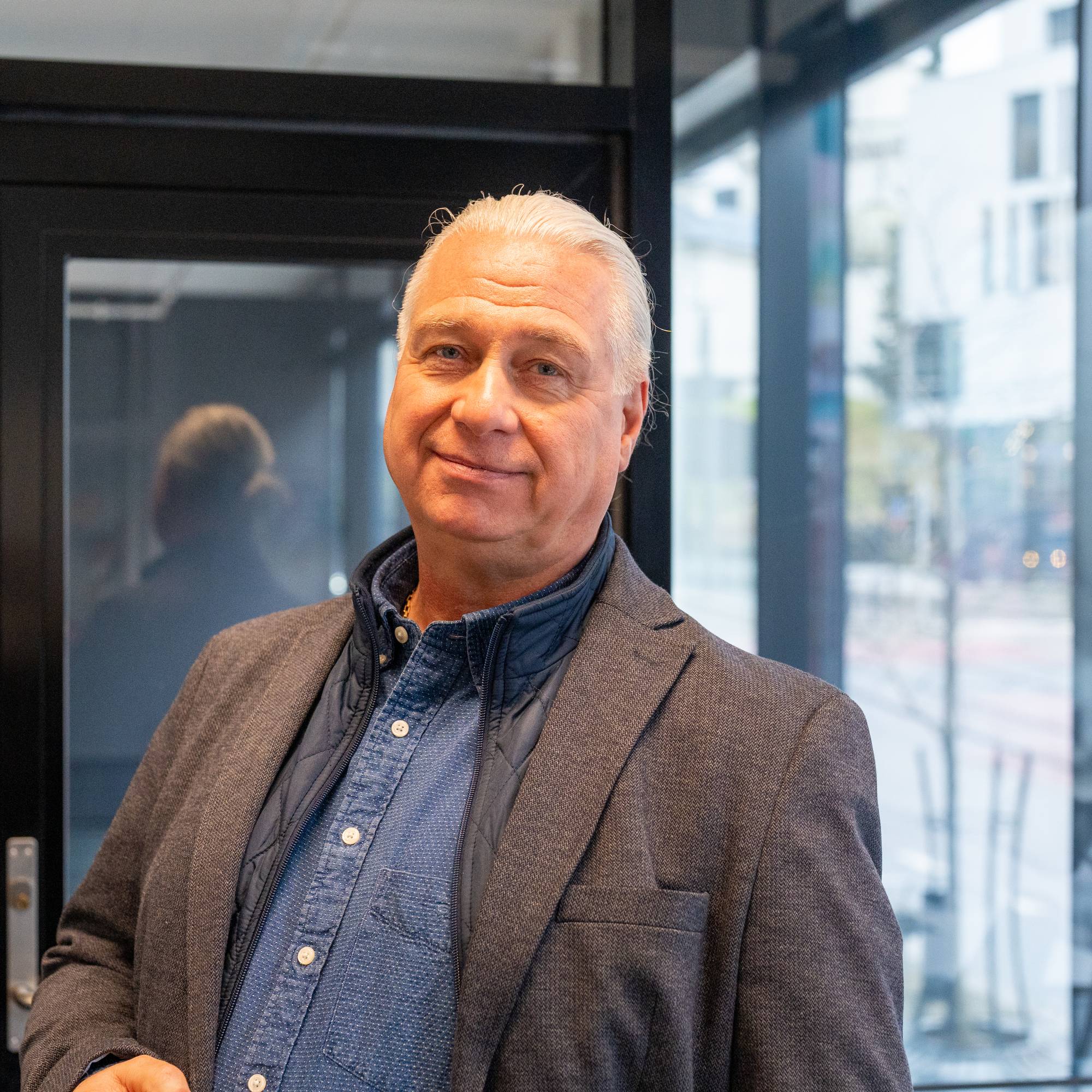 Portrett av en eldre mann med grått hår i blazer, stående innendørs ved en glassvegg. (Bildebeskrivelsen er laget av en KI-tjeneste)