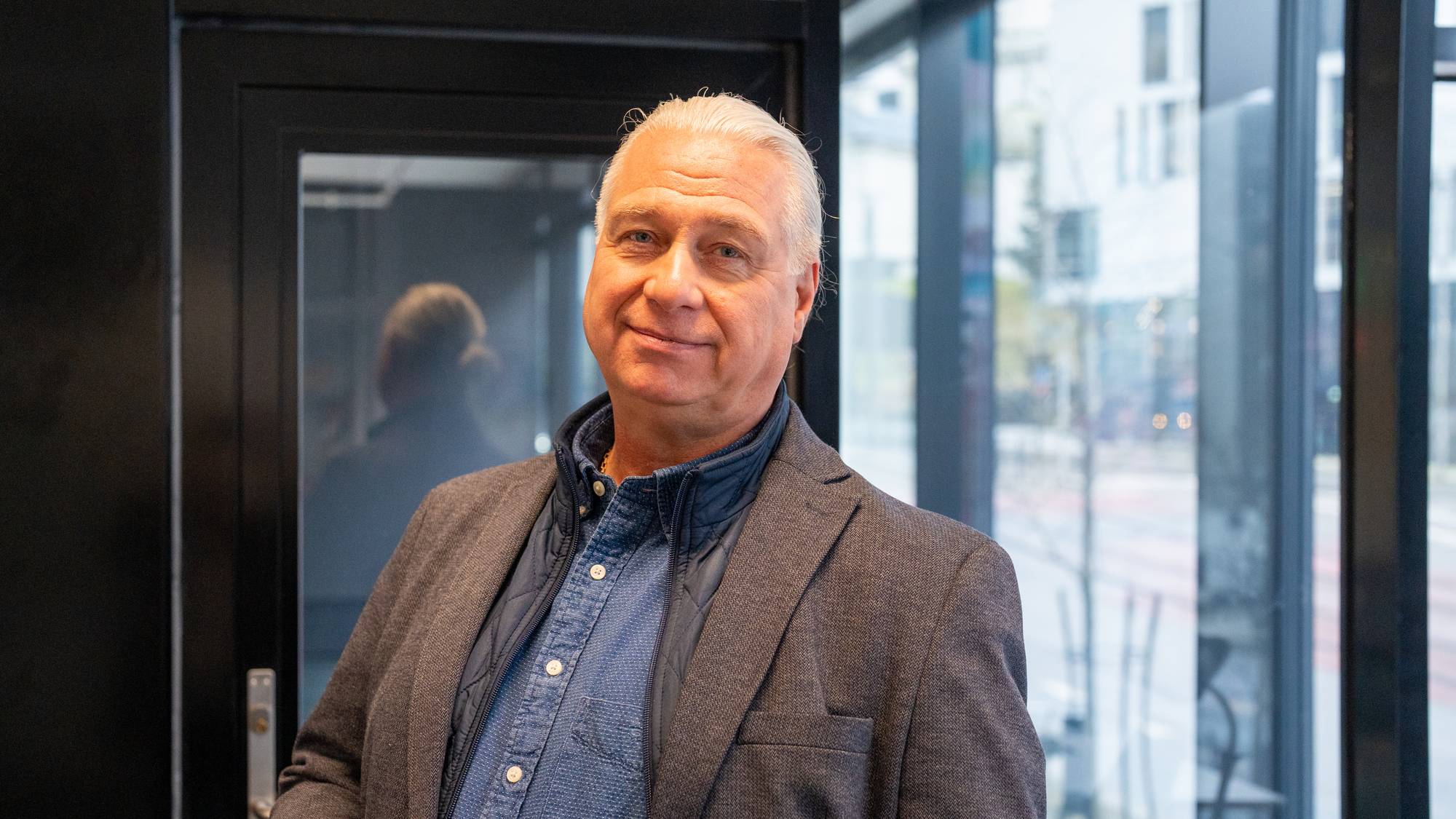 Portrett av en eldre mann med grått hår i blazer, stående innendørs ved en glassvegg. (Bildebeskrivelsen er laget av en KI-tjeneste)