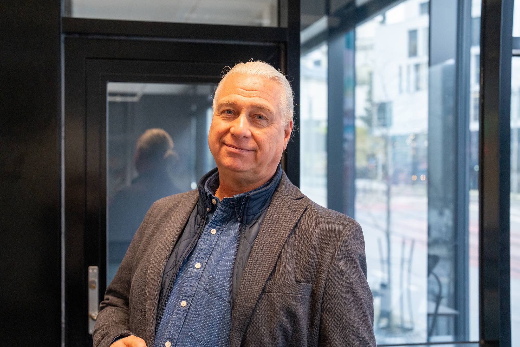 Portrett av en eldre mann med grått hår i blazer, stående innendørs ved en glassvegg. (Bildebeskrivelsen er laget av en KI-tjeneste)